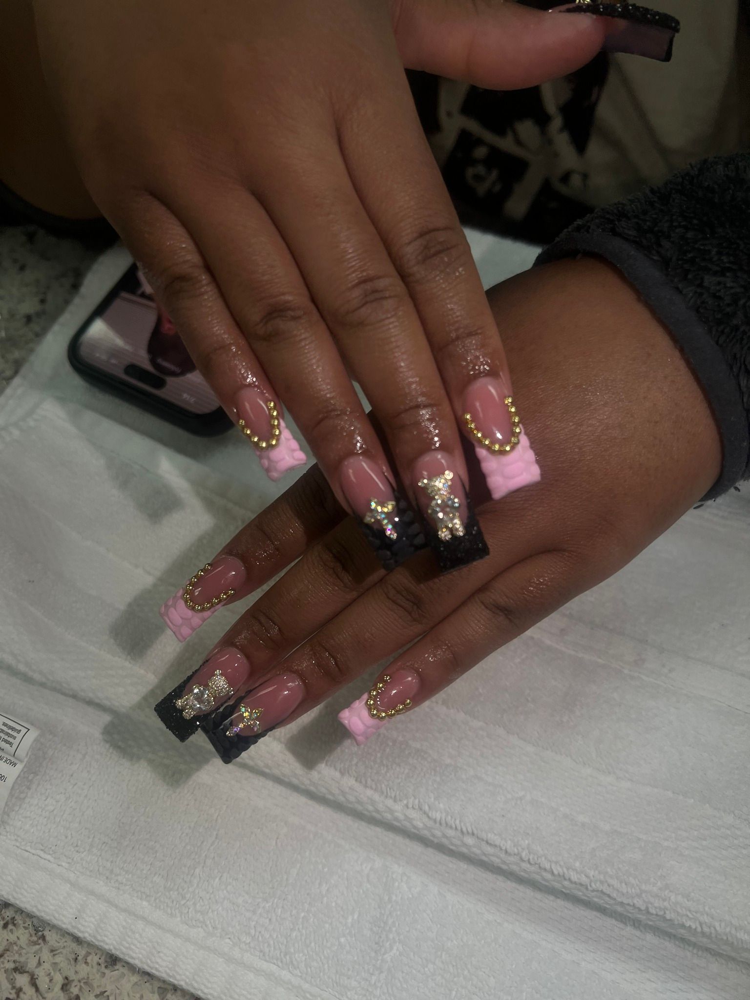 Hands with long square-shaped nails; black, pink, and gold design with rhinestones.