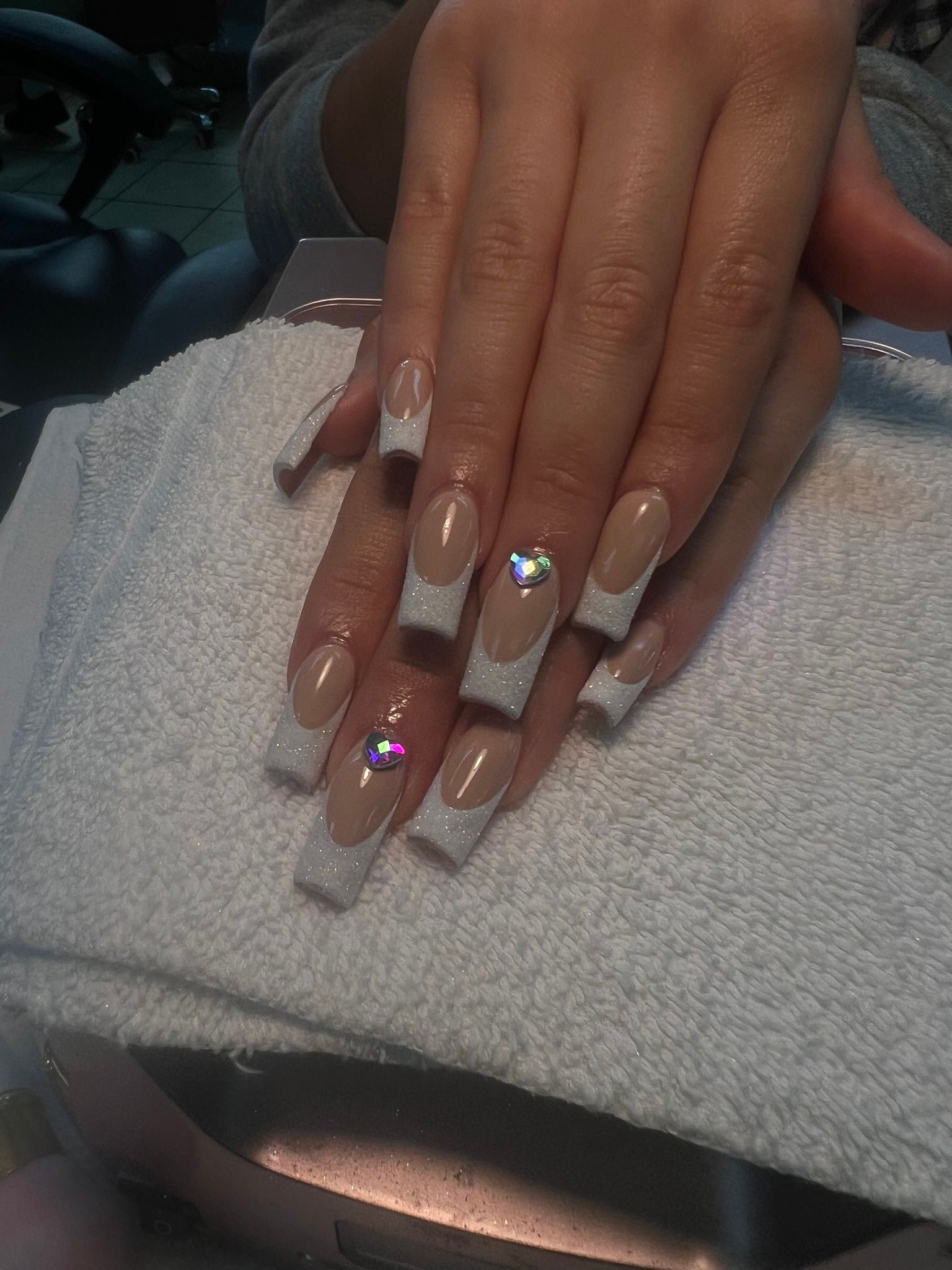 A hand with long, square-shaped French tip nails, adorned with glitter and rhinestones, rests on a white towel.