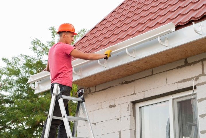 A construction worker in a hard hat on a ladder installing white gutters on the side of a brick house.