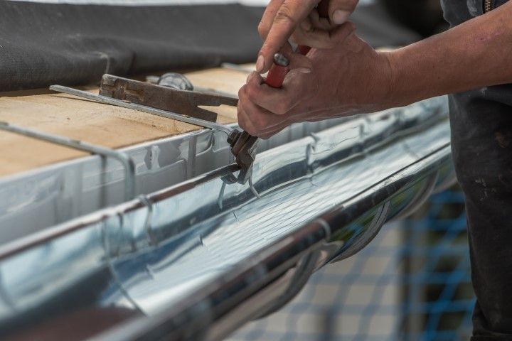 A person wearing red gloves uses snips to cut a piece of white, vented vinyl soffit while working on a ladder.