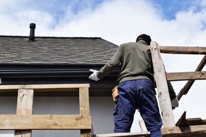 A construction worker stands on wooden scaffolding, reaching toward a dark roof gutter on a residential building.