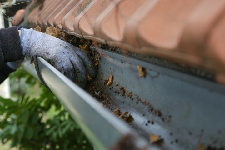 A person cleaning the gutter of a beige house under a clear blue sky.