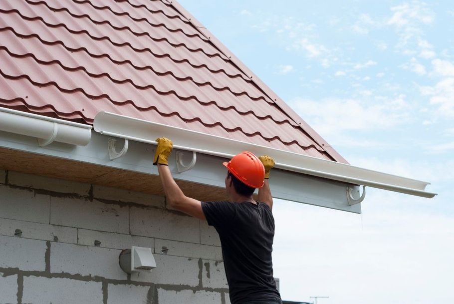 A worker in an orange hard hat and yellow gloves installs white gutters on the edge of a roof with red metal shingles.