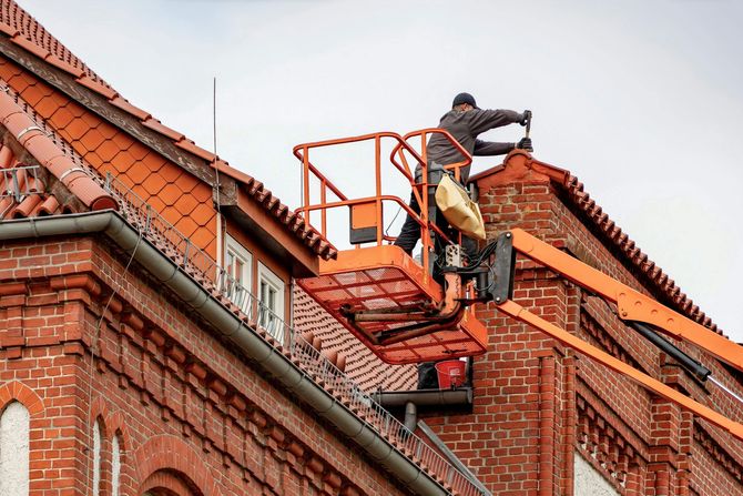A worker in an orange aerial lift repairs the roofline of a historic red brick building.