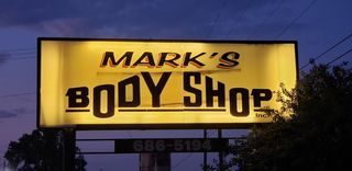 Sign for Mark's Body Shop, lit up with a yellow glow against a dusk sky.  Includes phone number below.