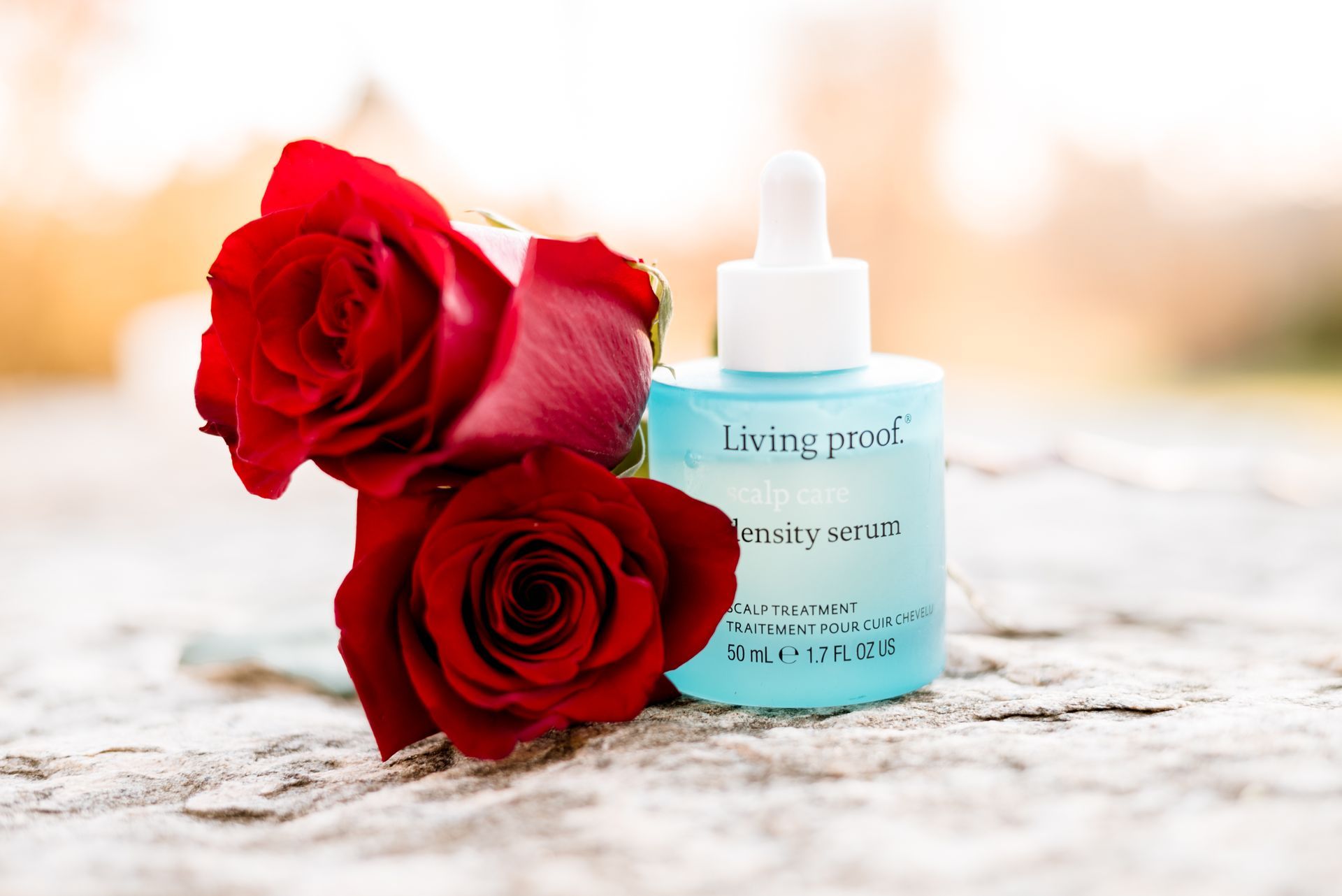 A blue bottle of Living Proof hair serum beside two red roses on a stone surface.