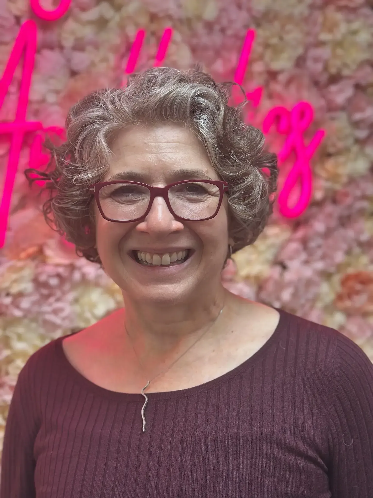 Woman with glasses smiles in front of a flower wall with neon pink text.