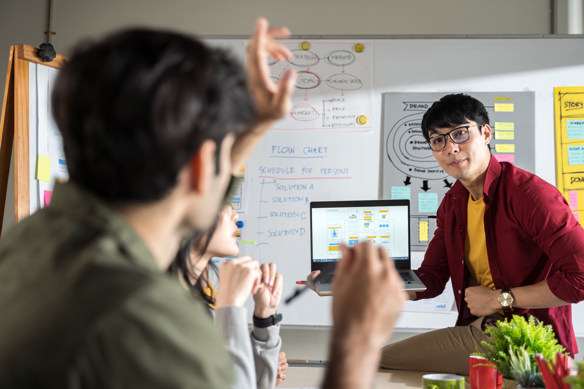 Man presenting laptop, pointing at screen, team in office setting with whiteboard and diagrams.
