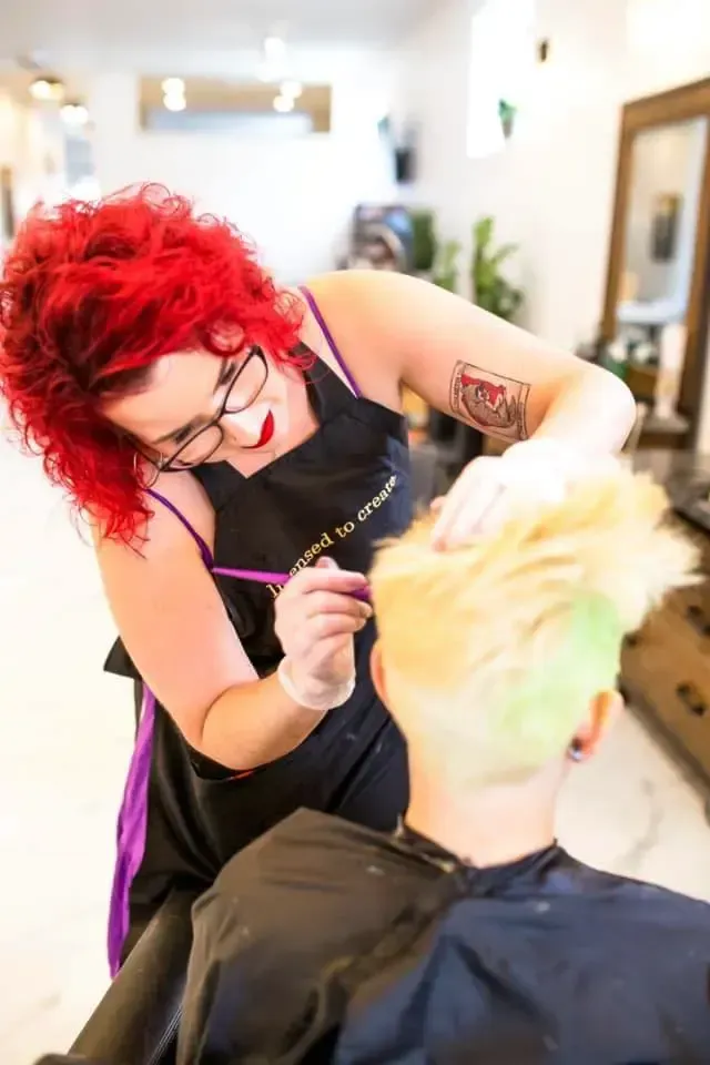 Hairdresser with red hair coloring client's blonde and green hair in a salon.