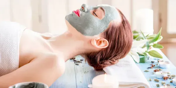 Woman with a facial mask relaxes at a spa, with towel, flowers, and candles in the background.