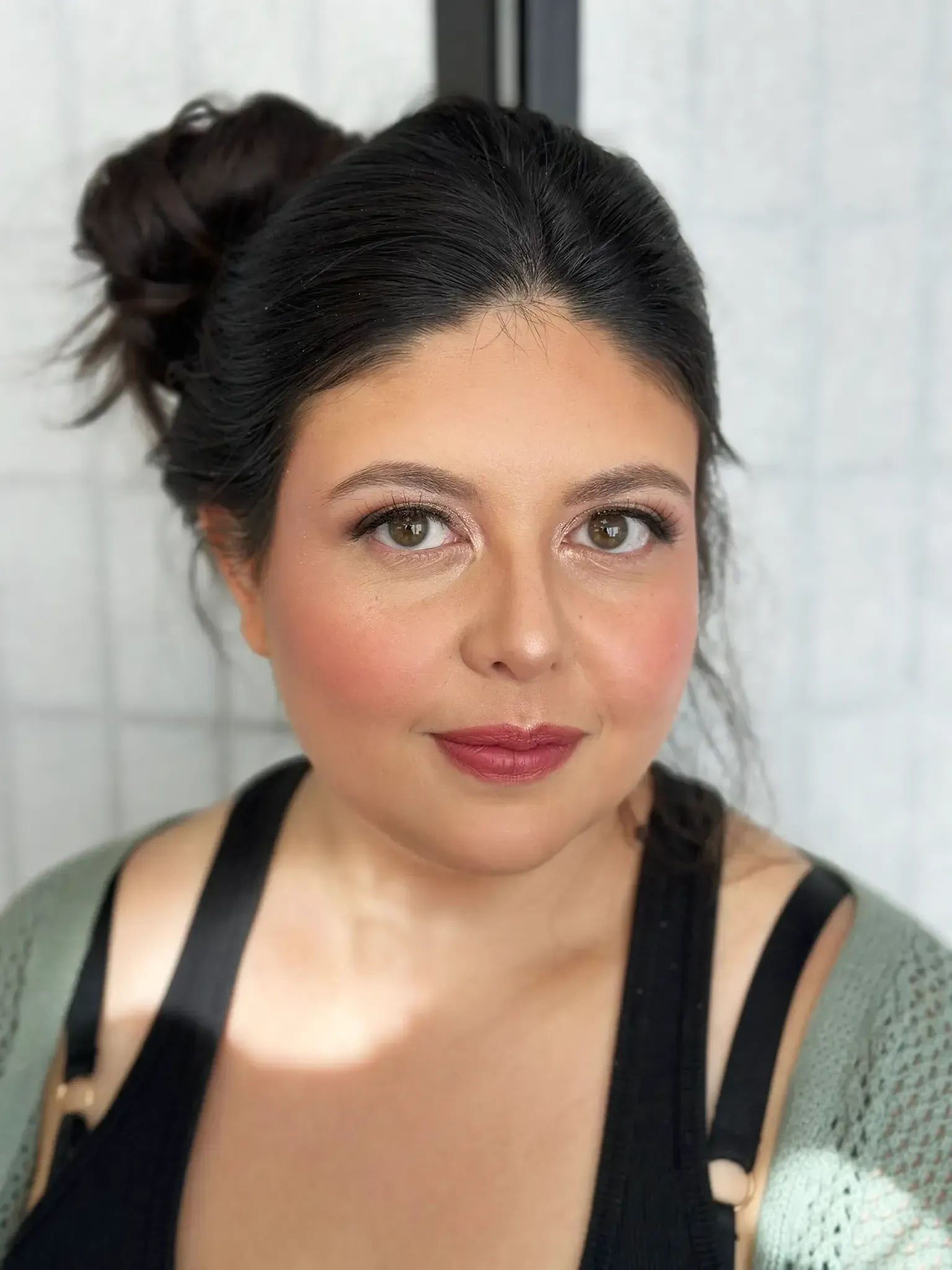 Woman with dark hair in a bun, wearing makeup, a black top, and green cardigan; lit by soft light.