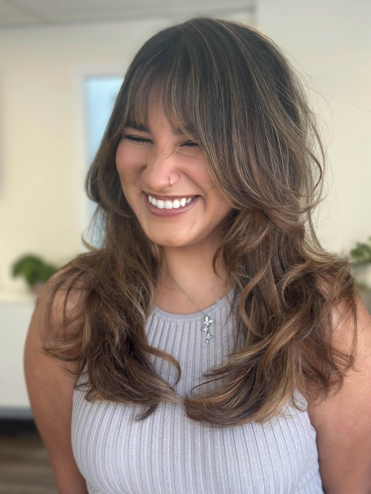 Woman with long, wavy brown hair and bangs smiles, wearing a gray tank top, indoors.