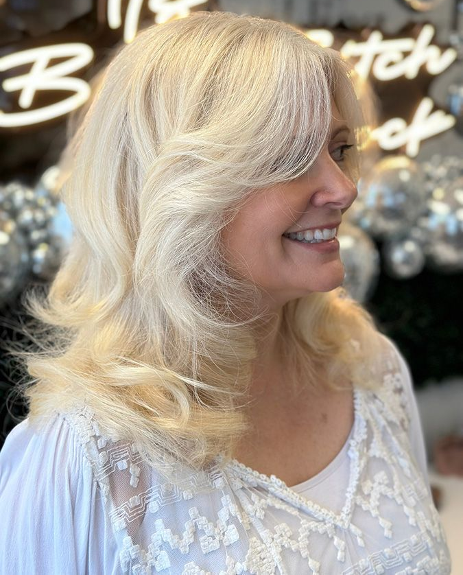 Woman with wavy, blonde hair, wearing a white, lace-trimmed top, smiling.
