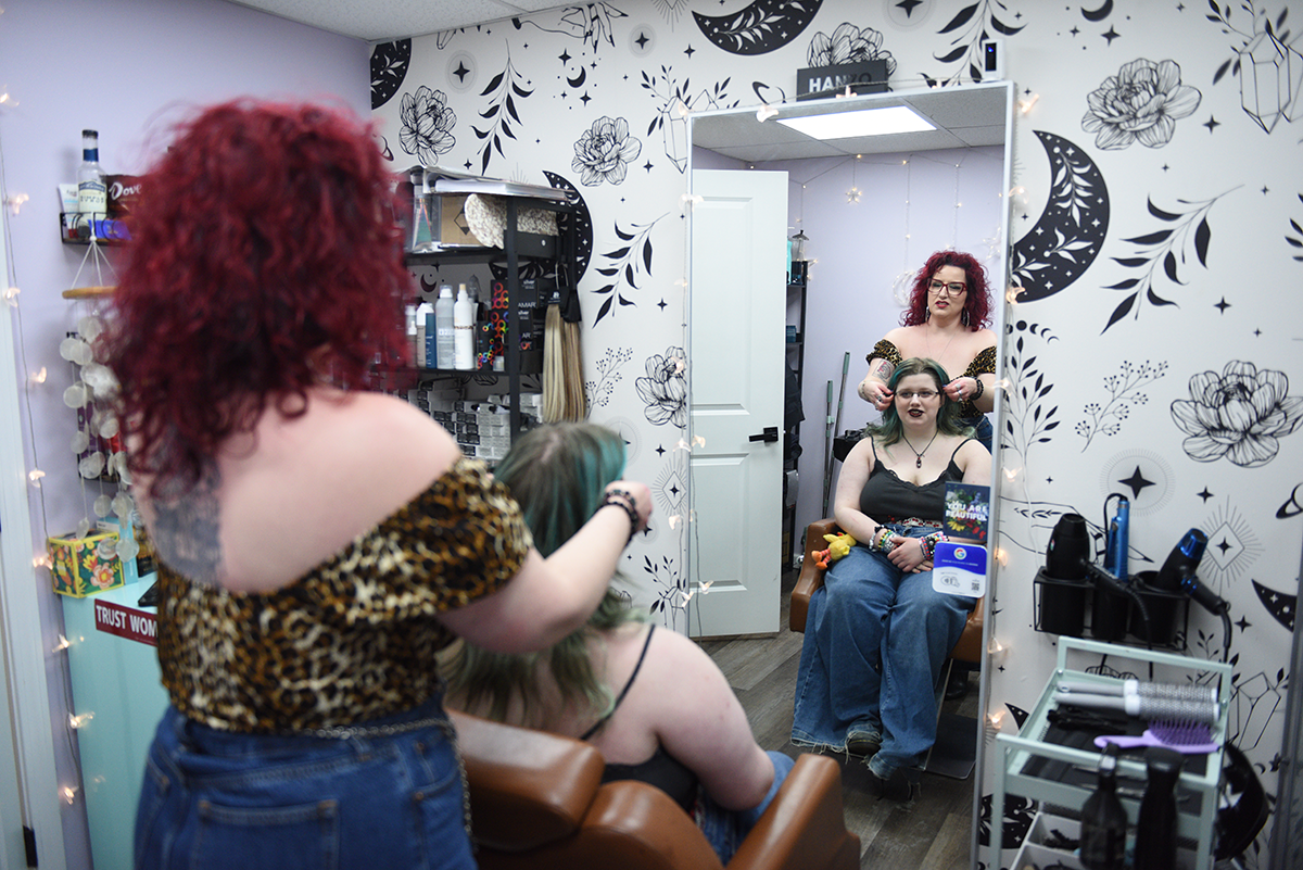 A hairstylist with red hair styles a client's hair in a salon with a mirror, a dark floral wall pattern, and other supplies.
