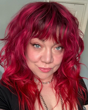 Woman with red, wavy hair and bangs, smiling, wearing a silver necklace, close-up.