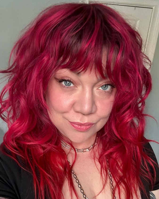 Woman with red, wavy hair and bangs, smiling, wearing a silver necklace, close-up.