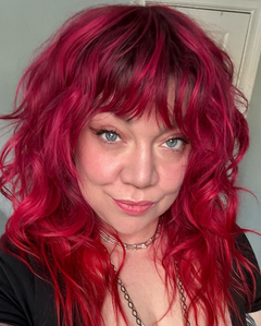 Woman with red wavy hair and bangs, smiling, wearing a necklace.