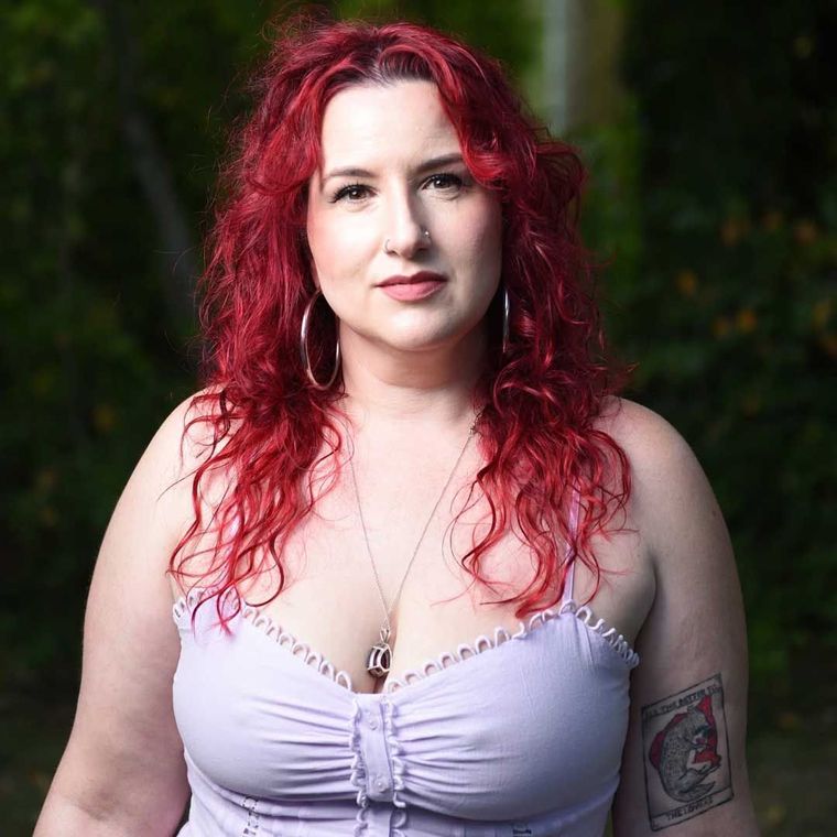 Woman with red curly hair in a lavender top. She has a tattoo and hoop earrings. Outdoors.