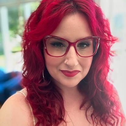 Woman with vibrant red hair, glasses, and lipstick smiles. Soft indoor lighting.