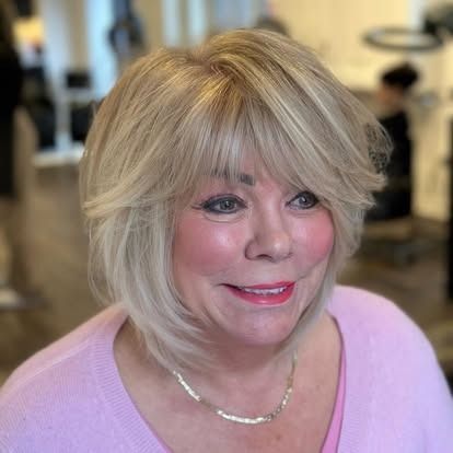 Woman with blonde layered bob and side-swept bangs, smiling, wearing a pink shirt, and a silver necklace.