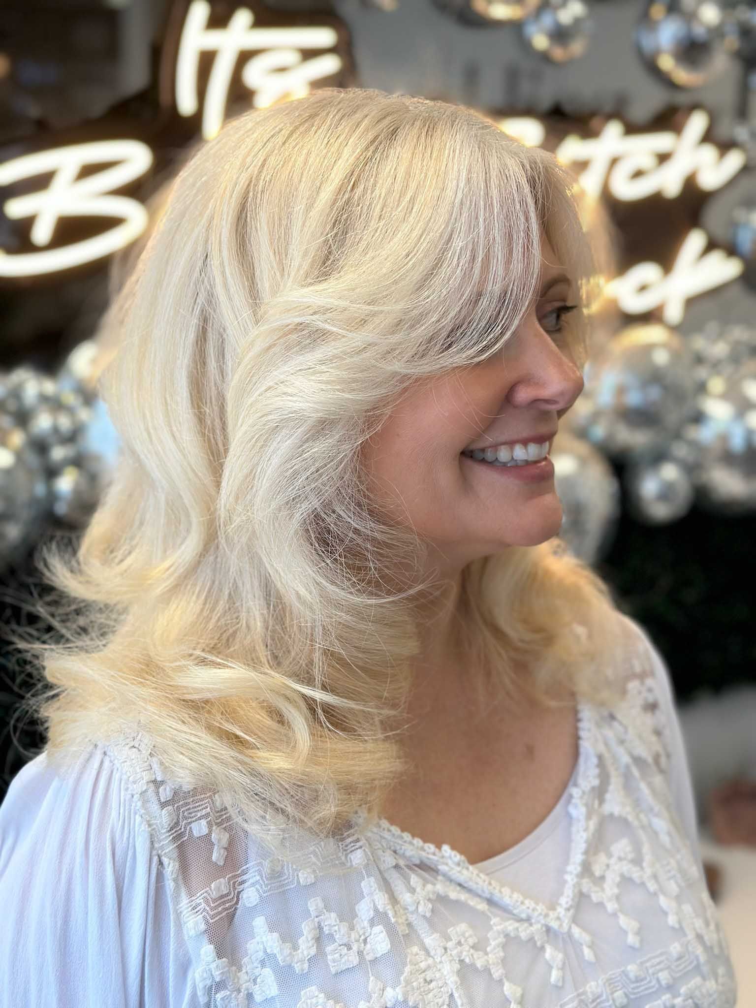 Blonde-haired person smiles, side profile. Wearing a white, lace top, and hair has side-swept bangs. 