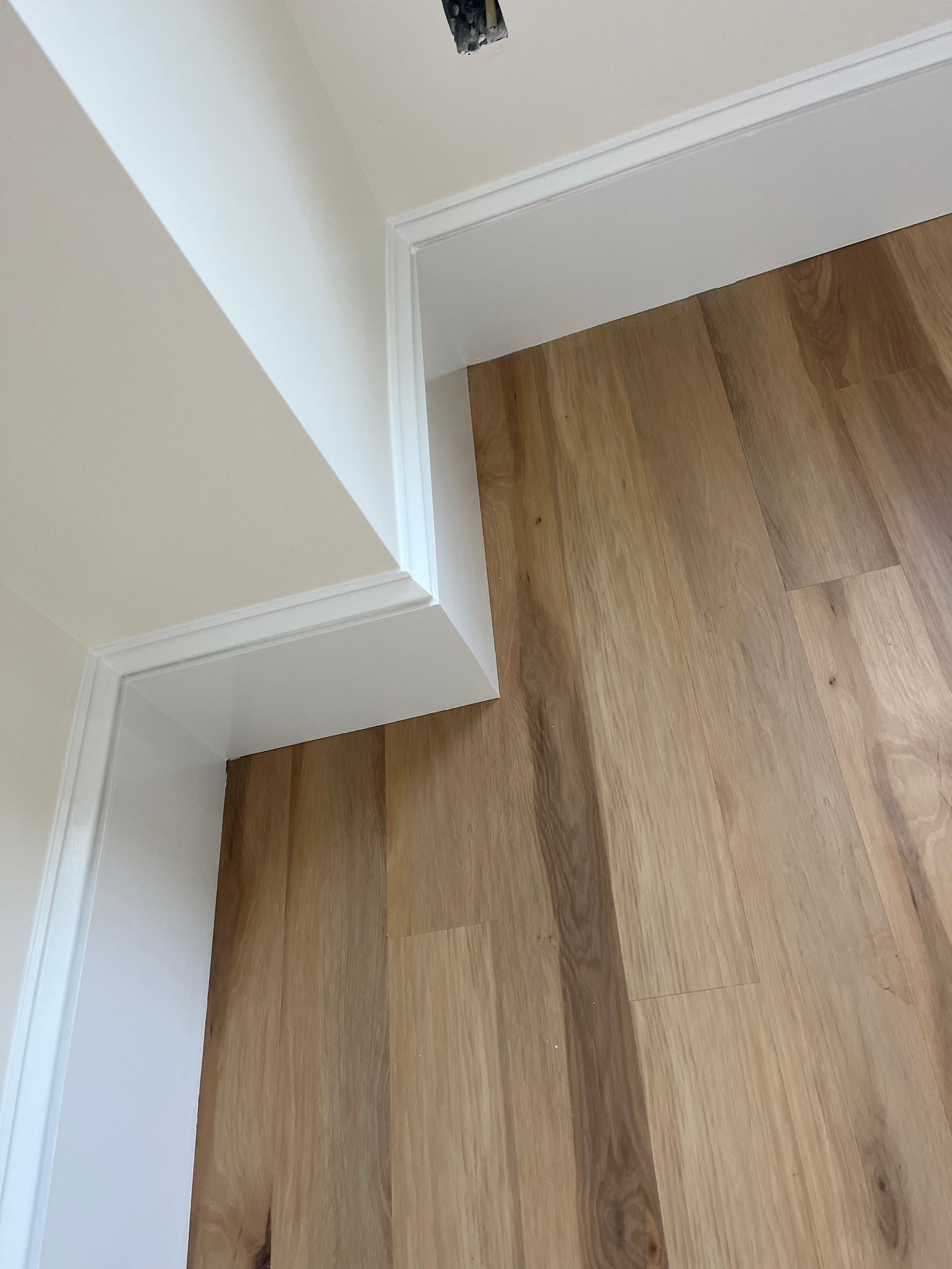 A high-angle view of a white corner wall with baseboards meeting light-colored wood flooring.
