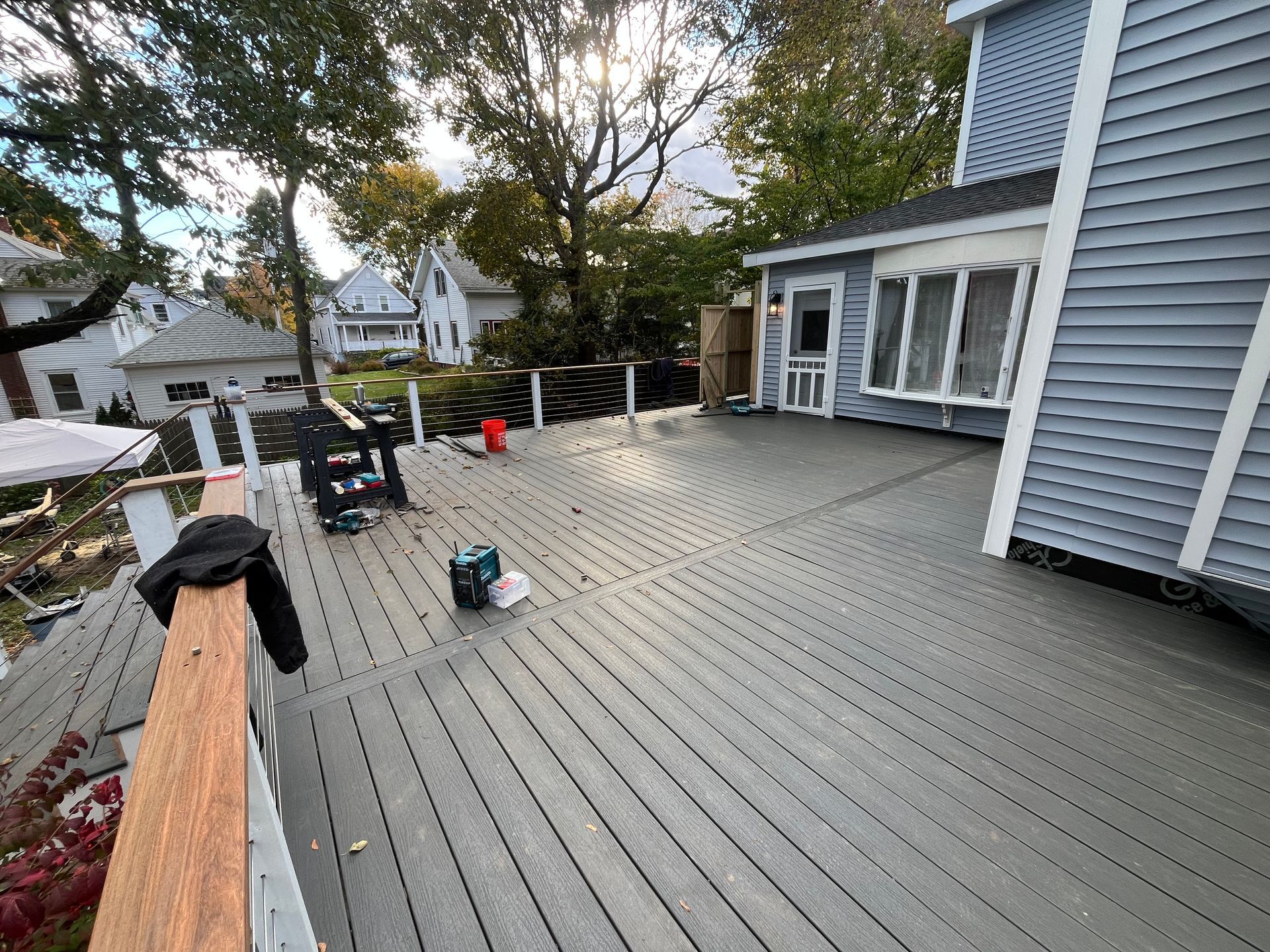A large grey deck with a white railing and a wooden handrail, viewed from above, with houses and trees in the background.