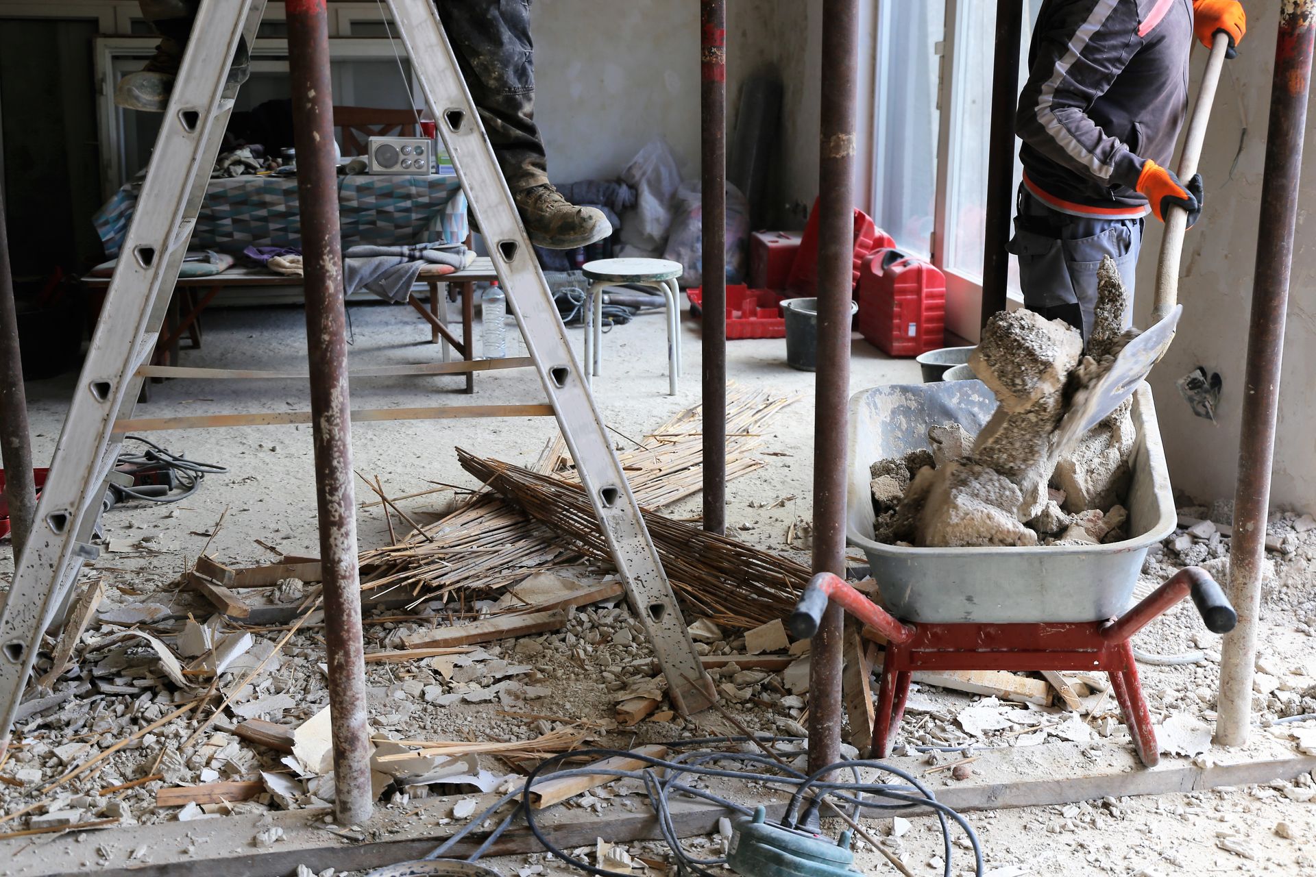 Construction workers clear debris from a renovation site using a shovel and a metal wheelbarrow amidst scaffolding.
