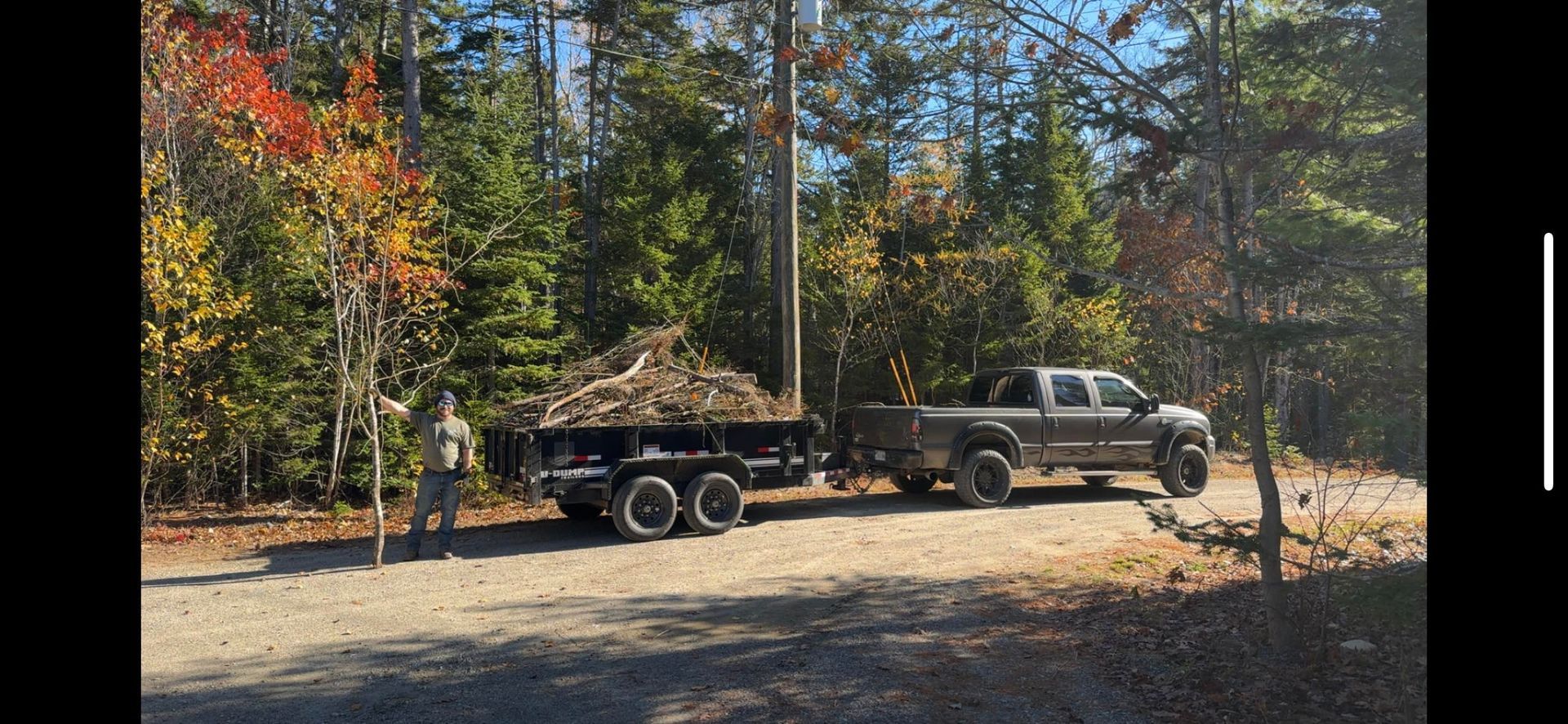 A person stands beside a pickup truck hauling a trailer filled with brush on a dirt road lined with autumn trees.