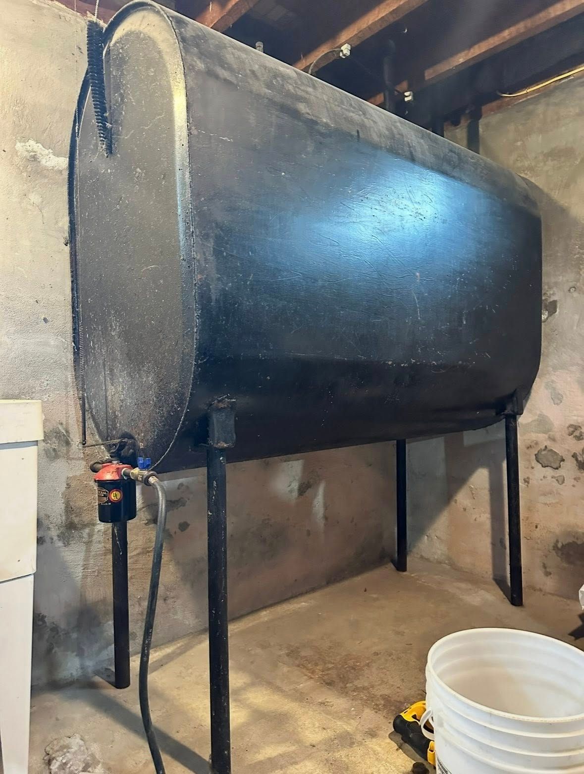 A black, oval oil storage tank on metal legs stands against a concrete basement wall with a bucket on the floor.