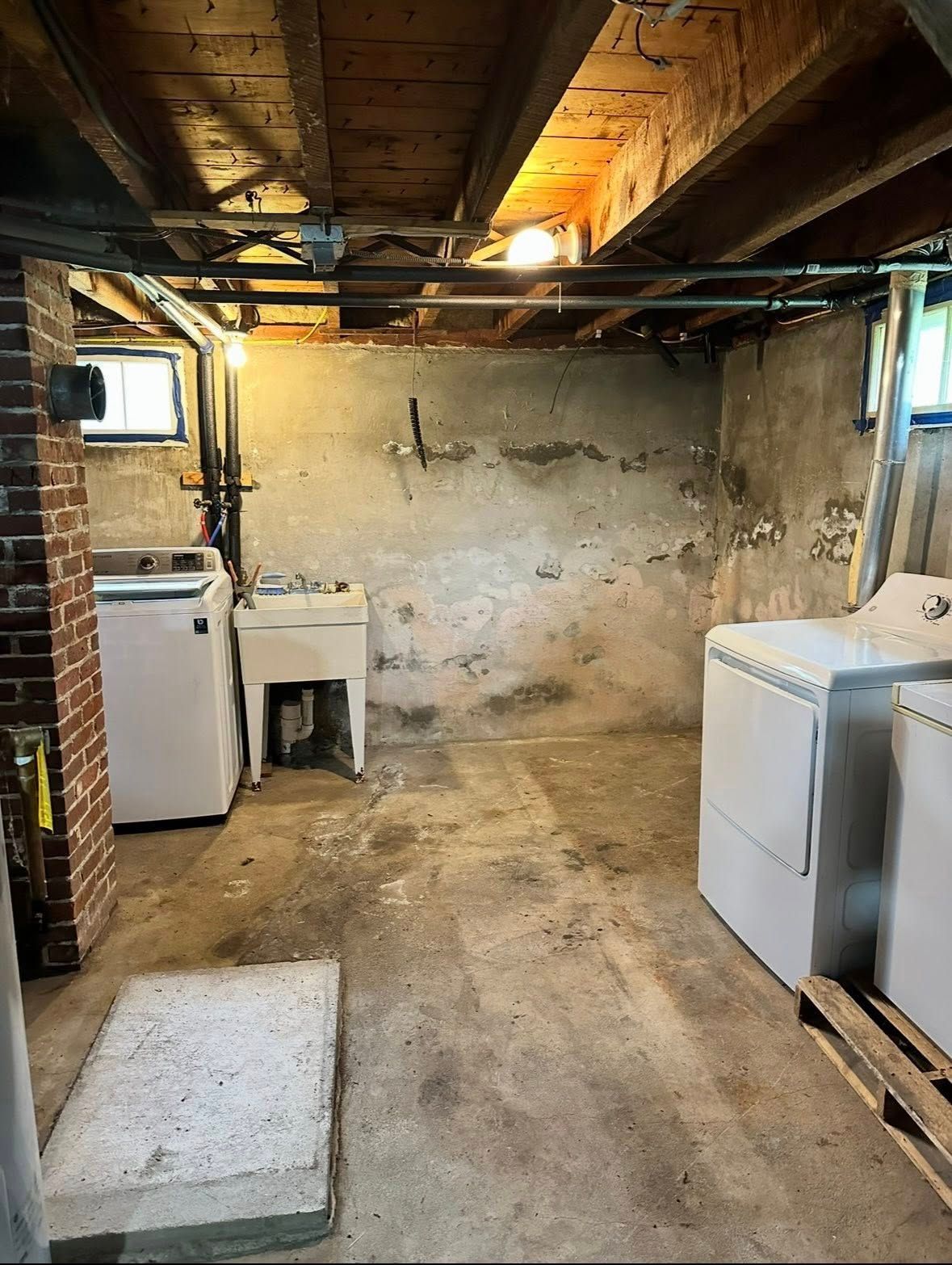 A basement laundry area with a white washing machine, utility sink, and dryer on a concrete floor beneath exposed joists.