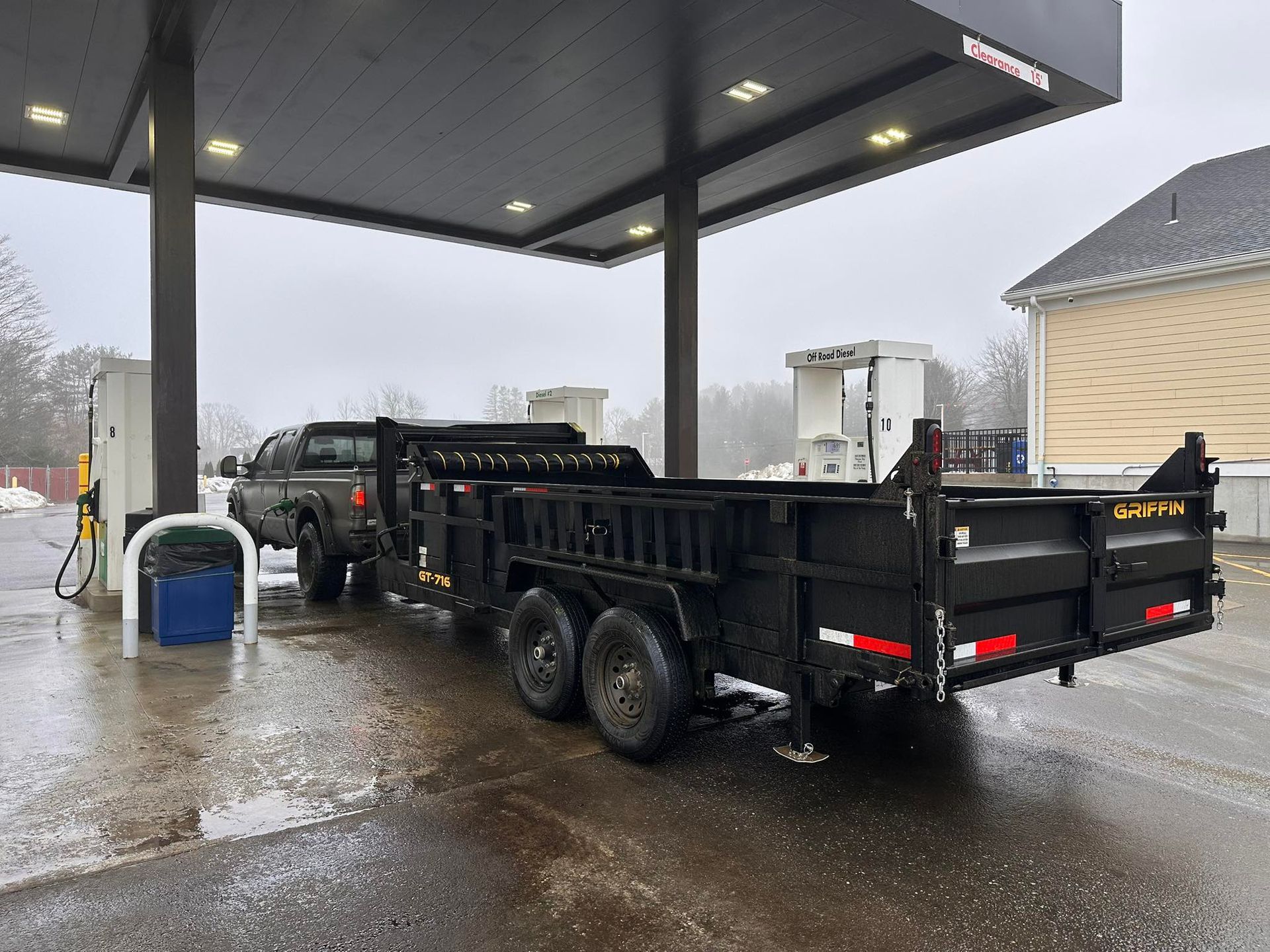 A dark-colored pickup truck pulling a large, black dump trailer parked under a gas station canopy on an overcast day.