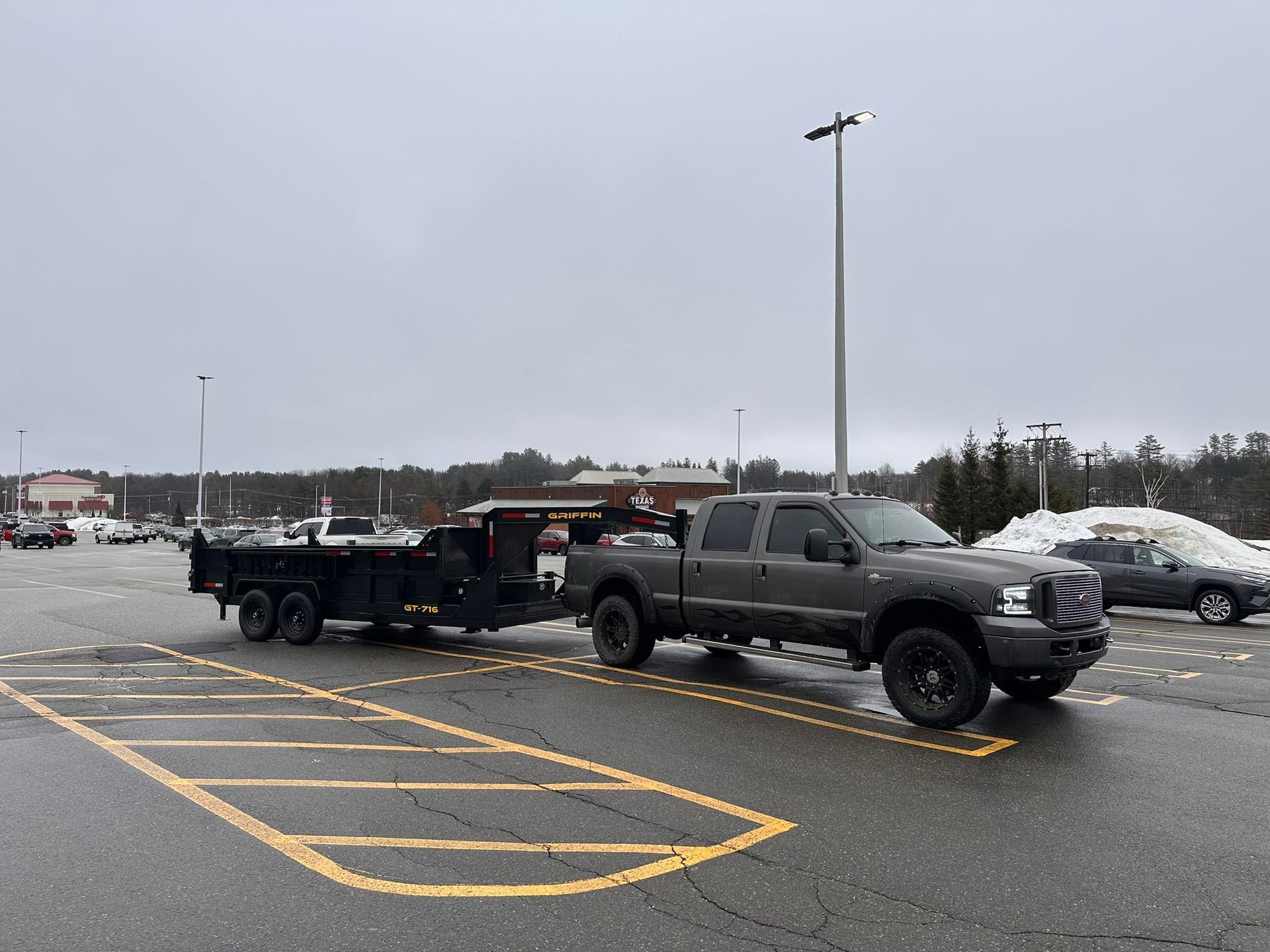 A dark-colored pickup truck towing a black flatbed trailer parked in an empty outdoor lot on an overcast day.