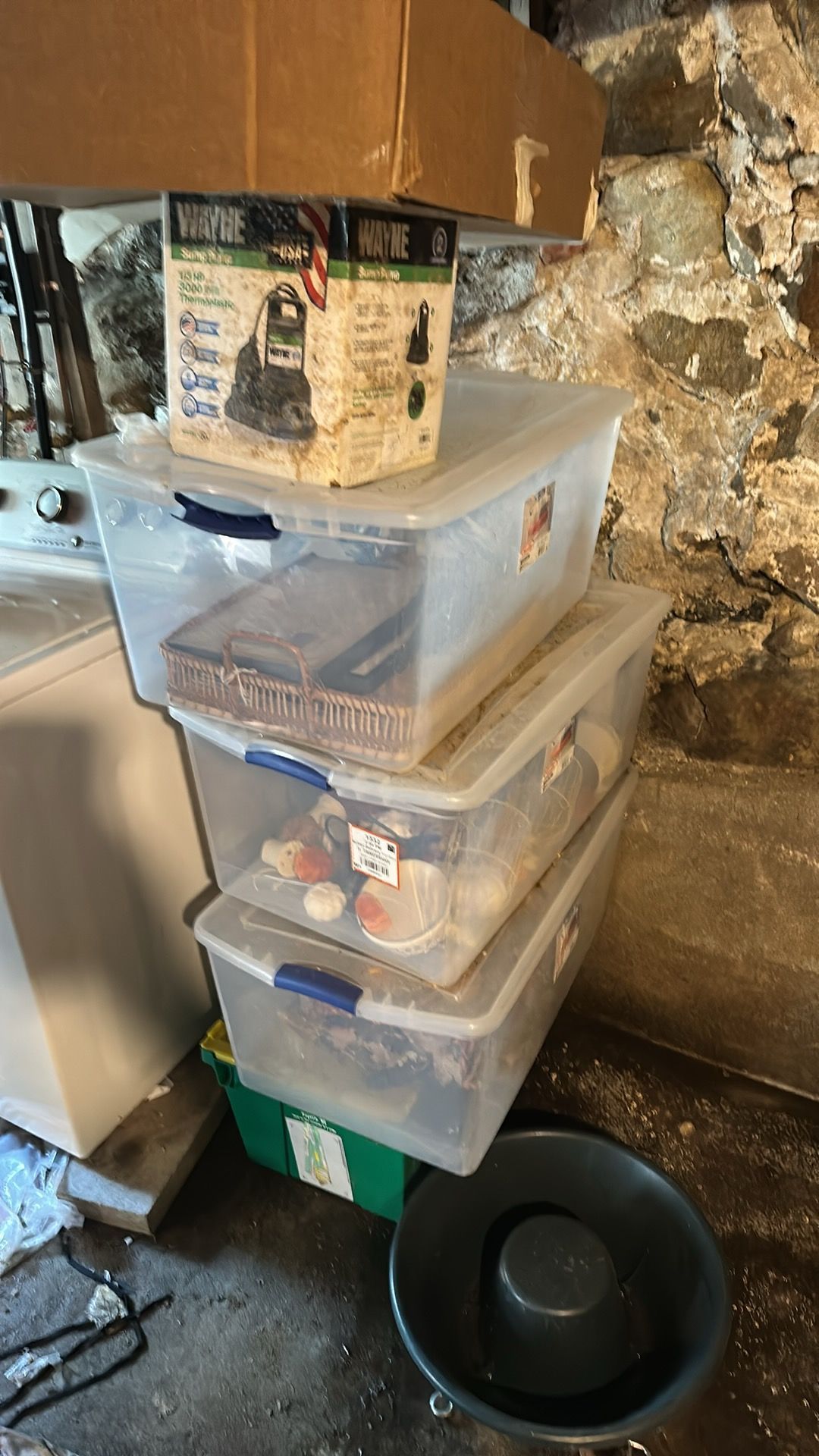 A stack of three clear plastic storage bins, a box, and a black bowl in a basement with a stone wall background.