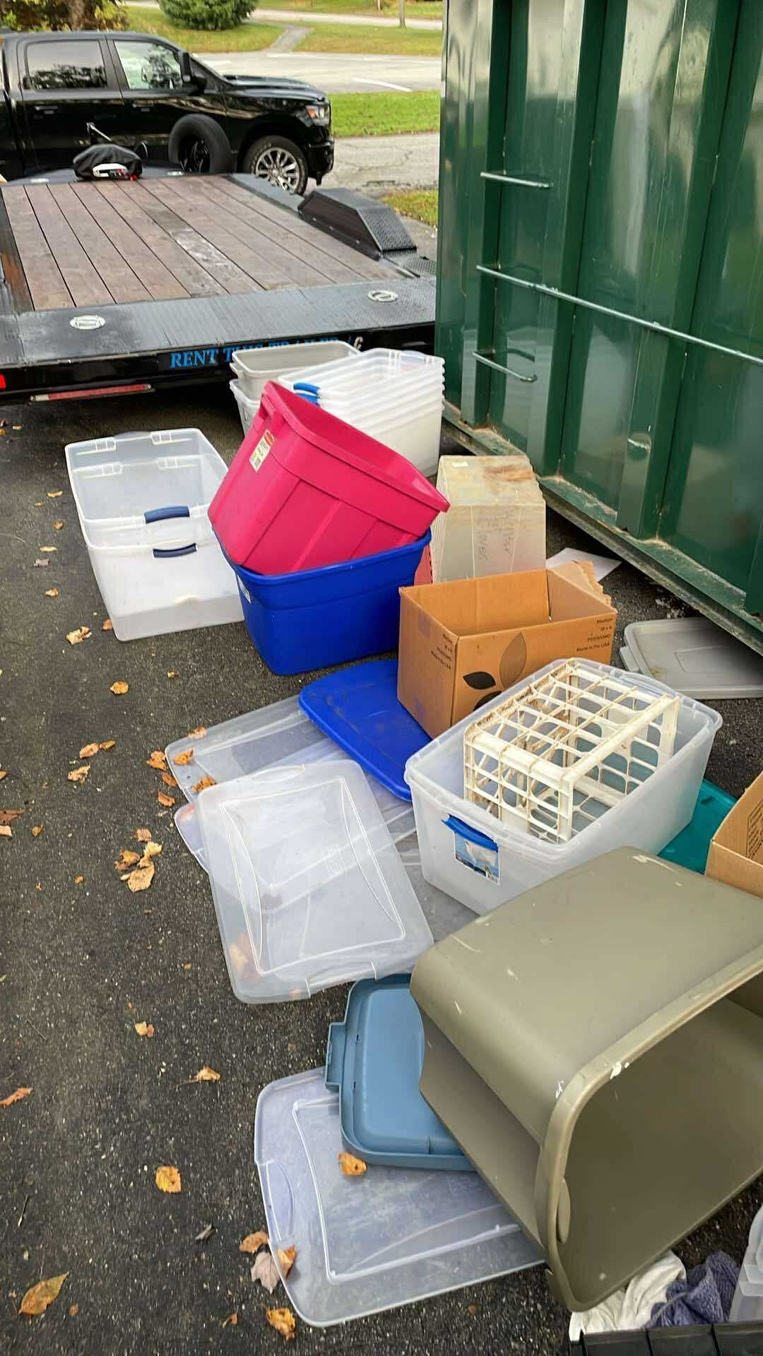 A collection of plastic storage bins and cardboard boxes piled on a driveway next to a large green dumpster and trailer.