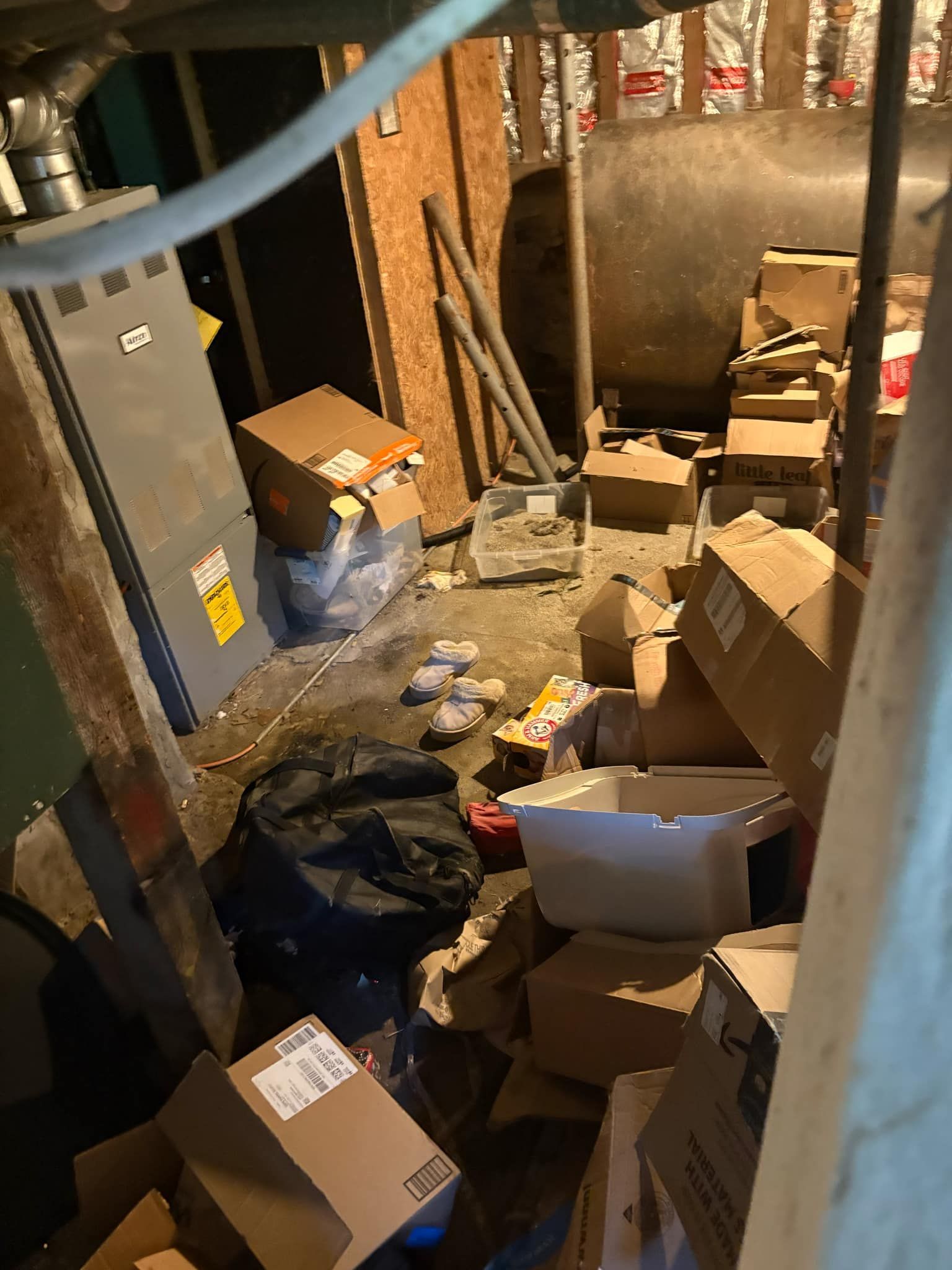 A basement area filled with cardboard boxes, a trash bag, and scattered items near a large furnace and fuel tank.