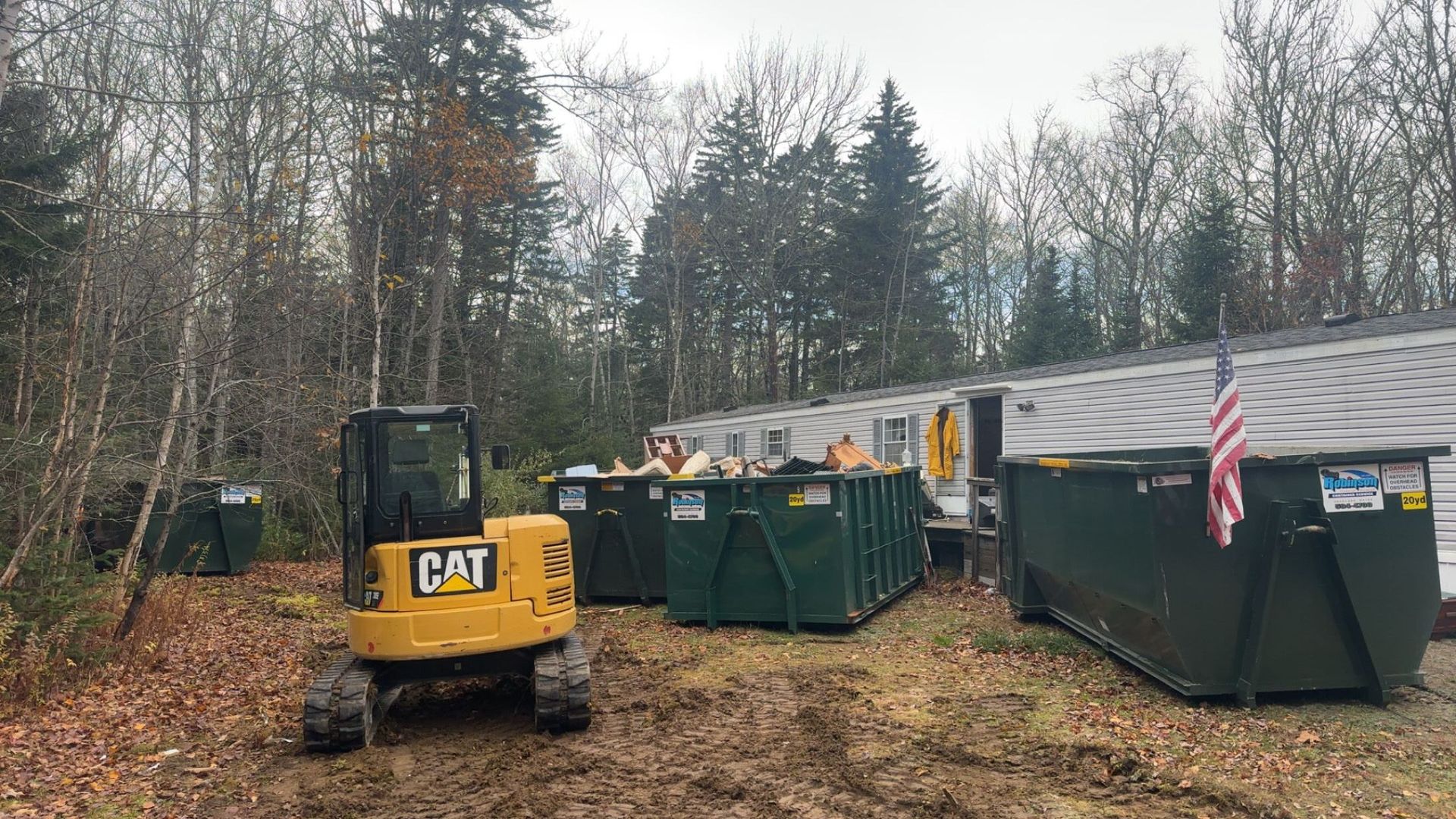 A yellow CAT excavator parked next to several green dumpsters filled with debris near a building with an American flag.