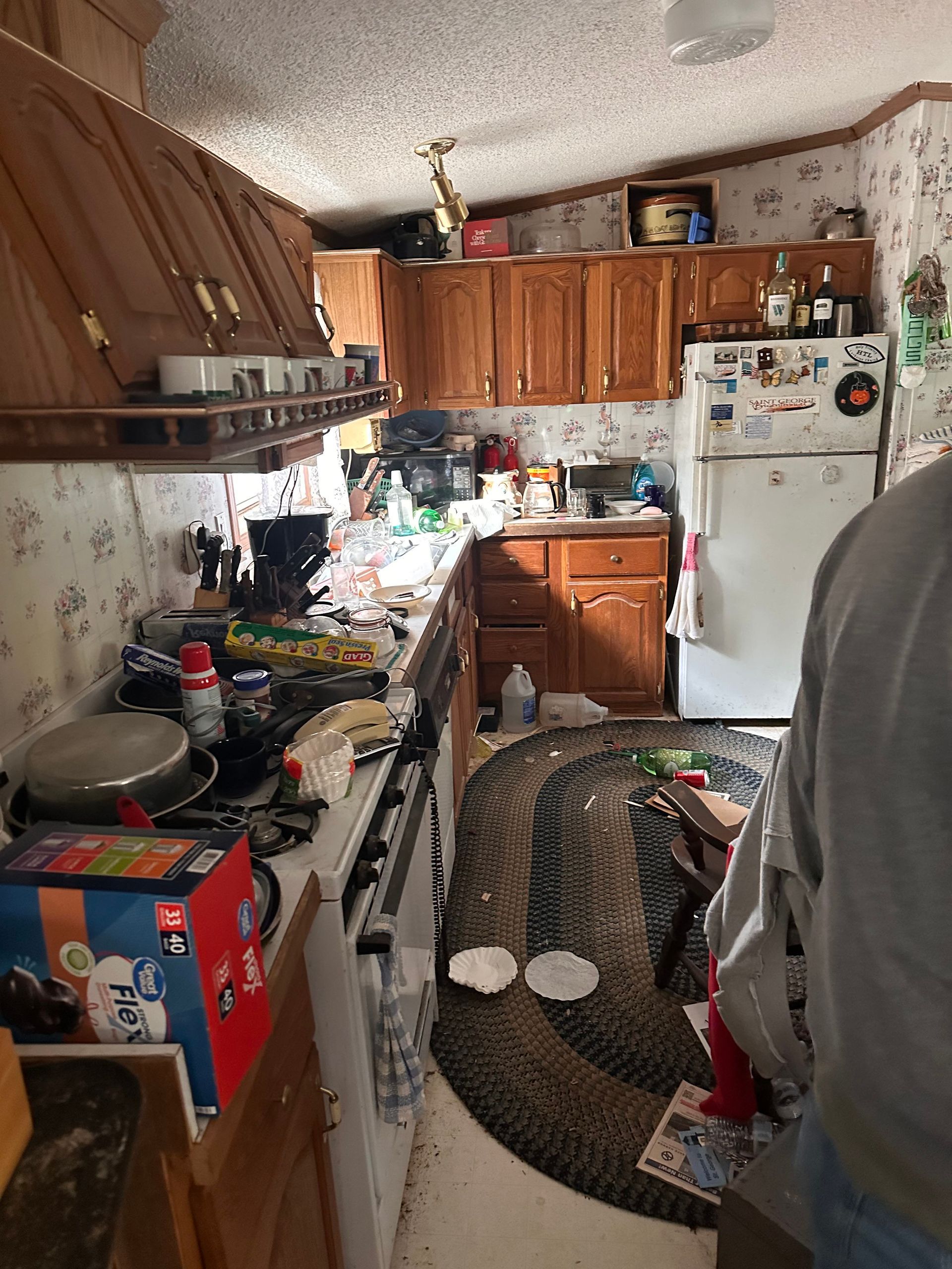 A cluttered kitchen with wooden cabinets, a white refrigerator, a stove, and a braided rug on the floor.