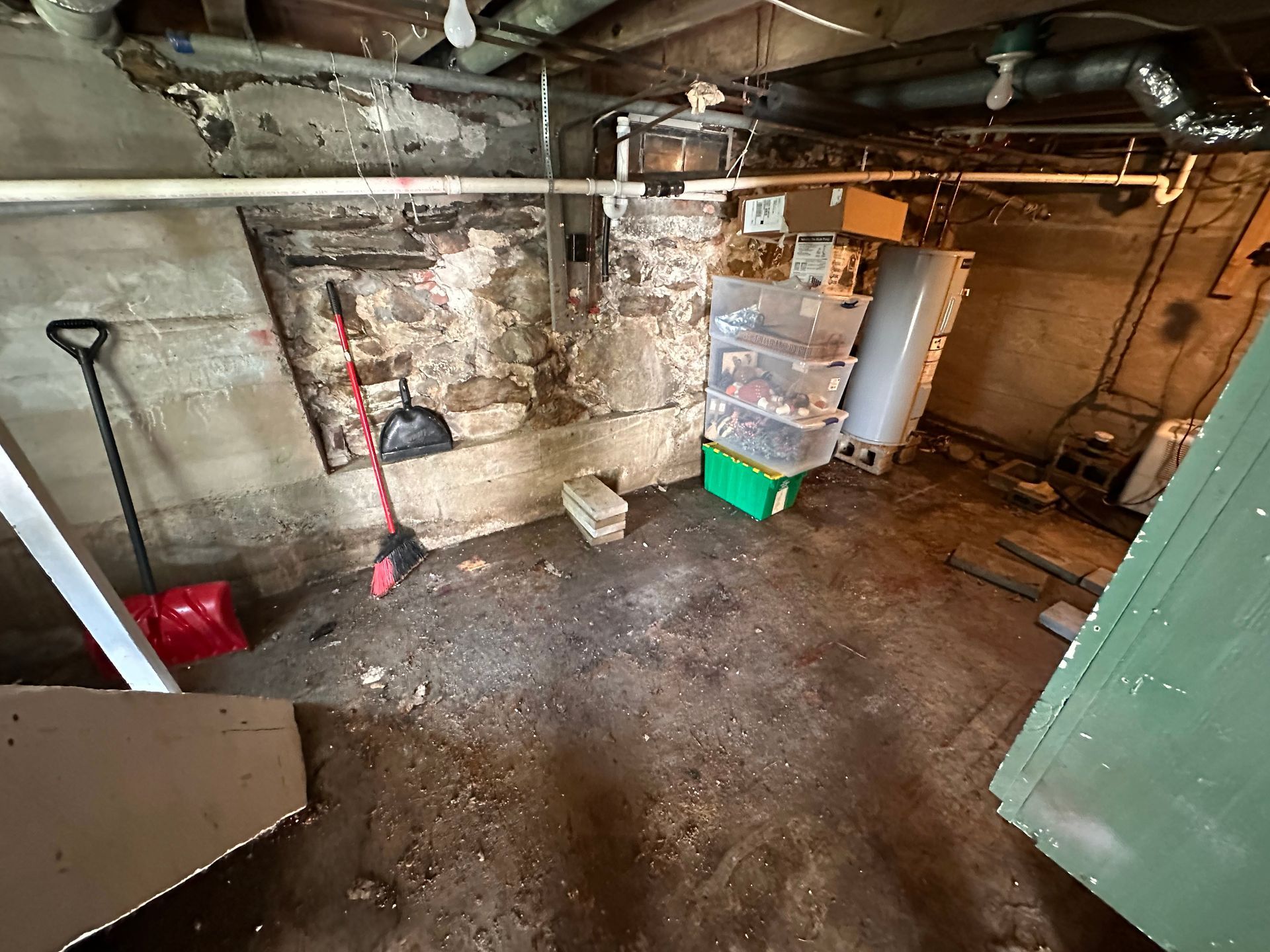 A dimly lit, unfinished basement with stone walls, a dirt floor, cleaning tools, a water heater, and storage bins.