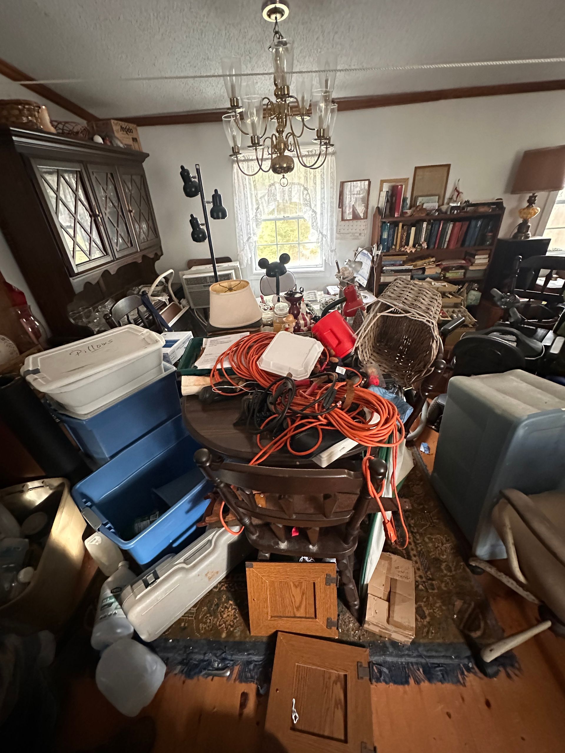 A cluttered dining room with a chandelier, wooden furniture, stacks of bins, and scattered orange extension cords.