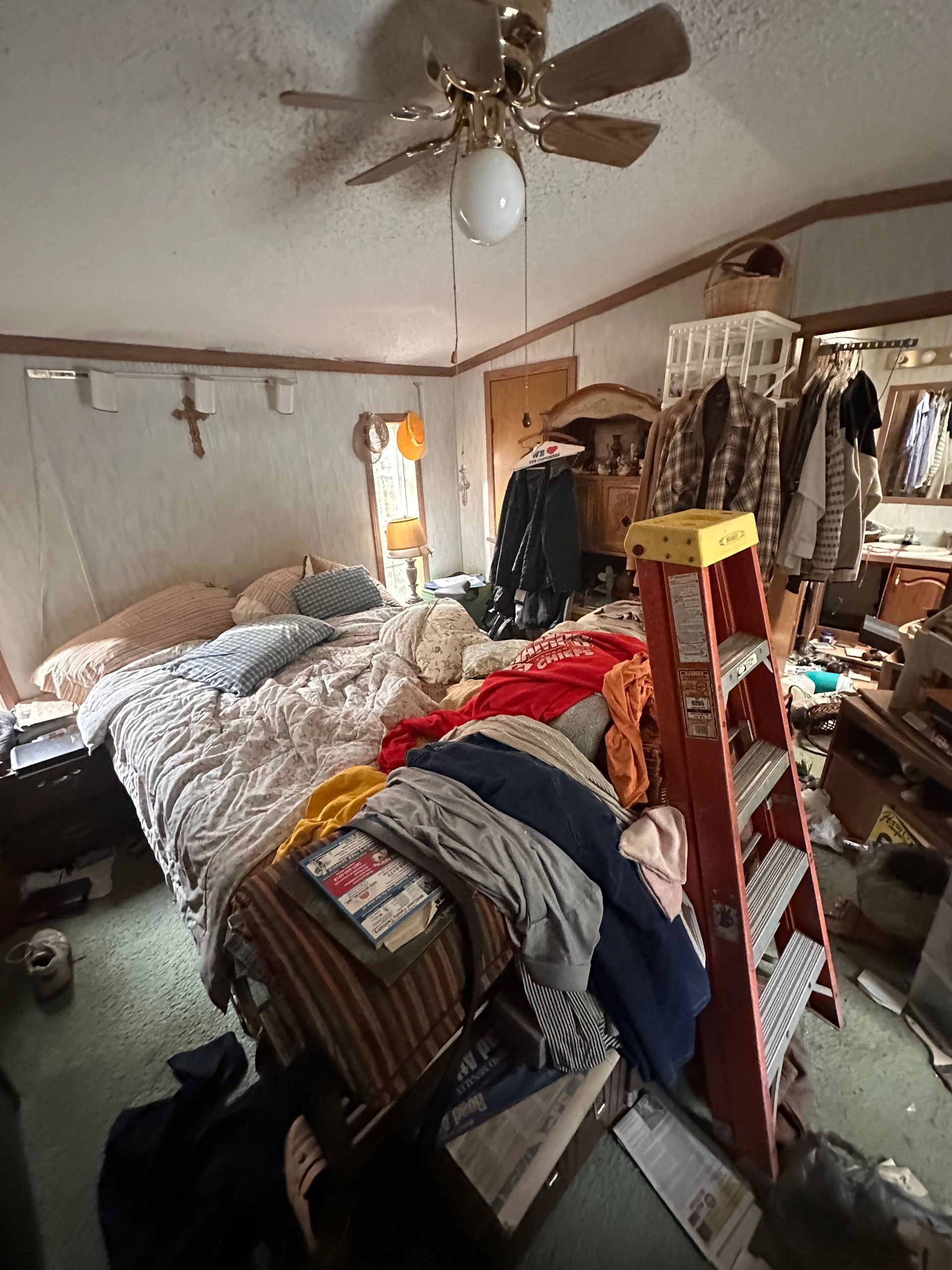 A cluttered bedroom featuring an unmade bed, a large orange ladder, and clothes hanging on racks in a lived-in space.