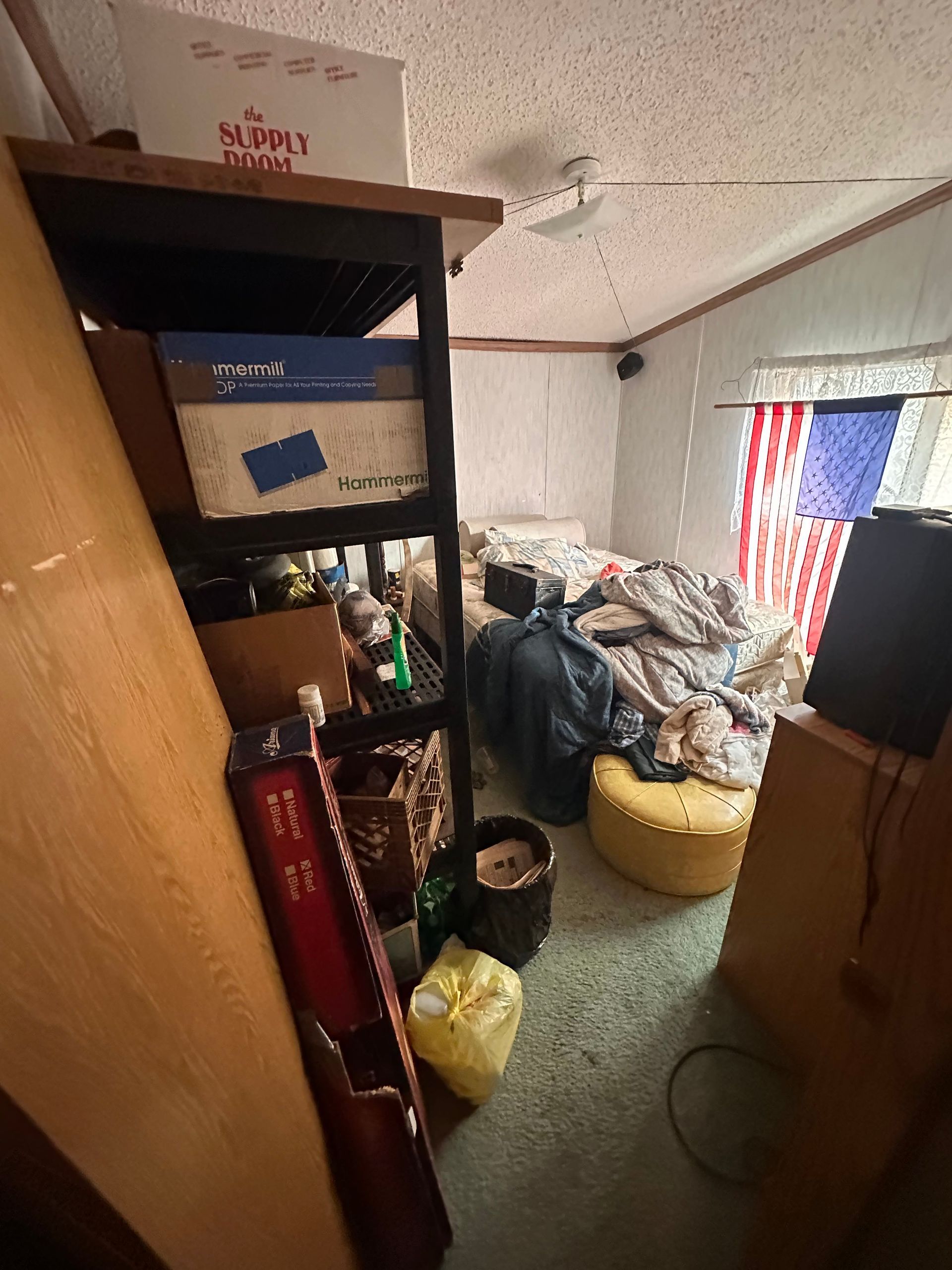 A messy bedroom with a bed, shelving unit filled with boxes, a yellow ottoman, and an American flag hanging in the window.