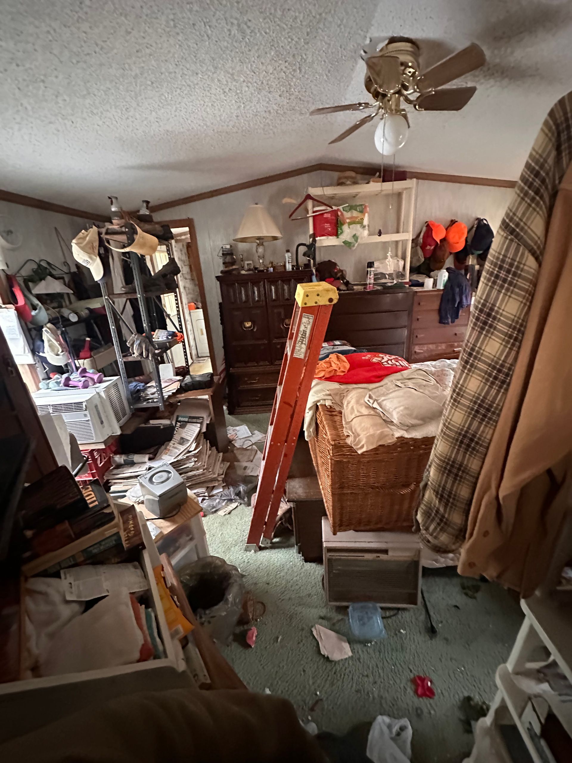 A cluttered bedroom with a ceiling fan, a tall wooden dresser, an orange step ladder, and miscellaneous items on the floor.