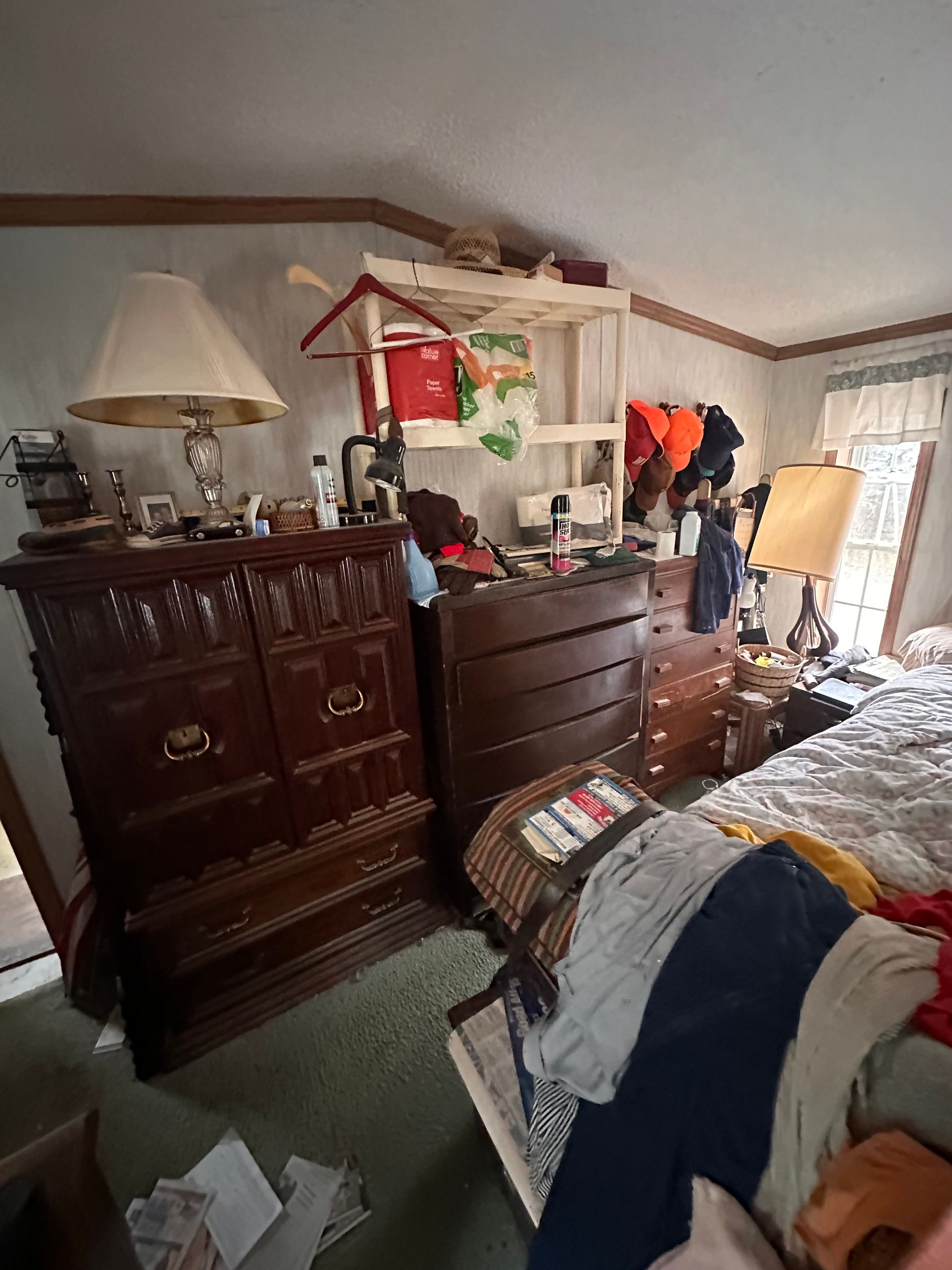 A bedroom with several dark wood dressers, a lamp, and a clothing rack against a wall, with laundry piled on a bed.