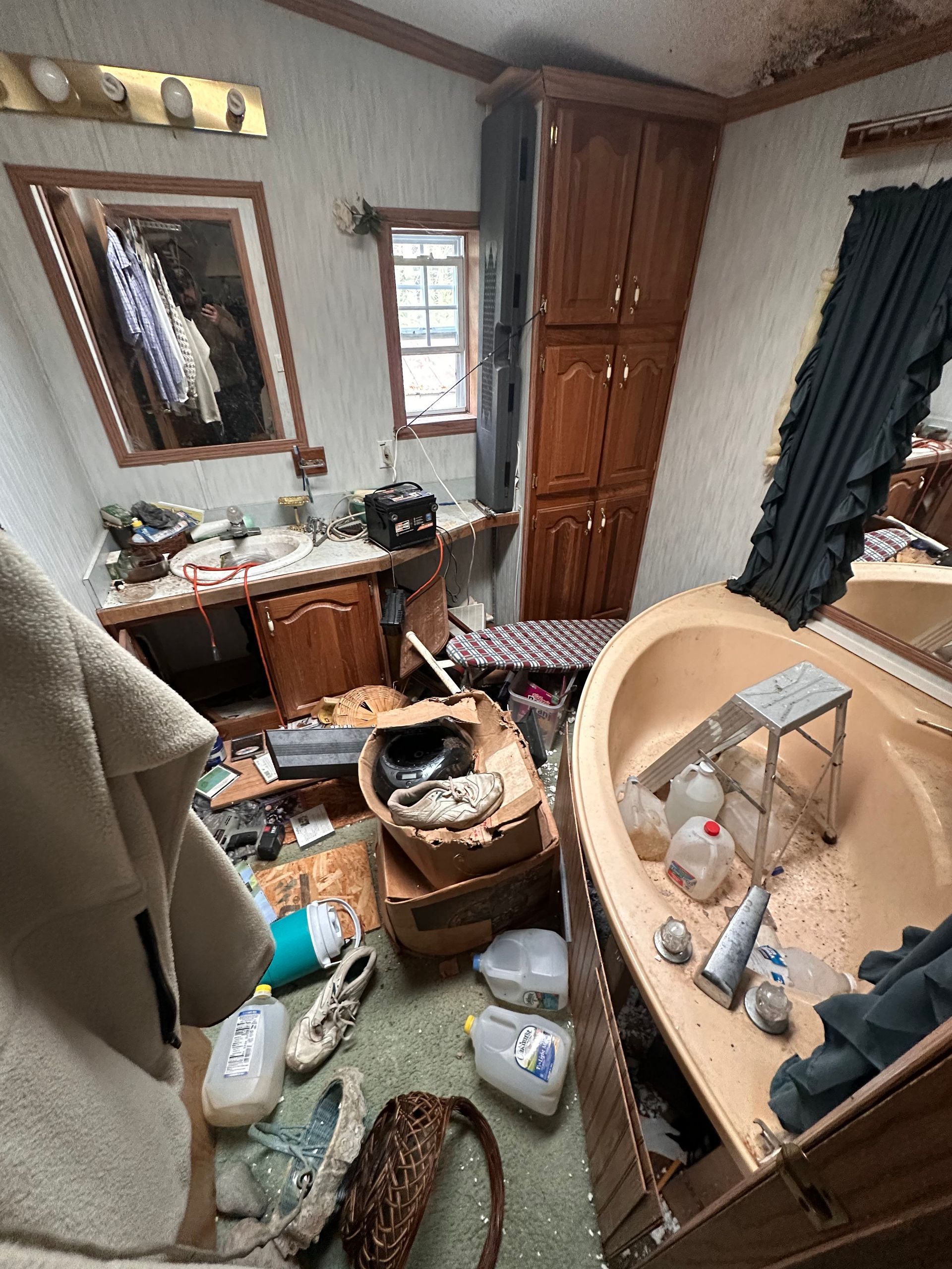 A cluttered bathroom featuring a beige bathtub, wood cabinets, a mirror, and various items scattered across the floor.