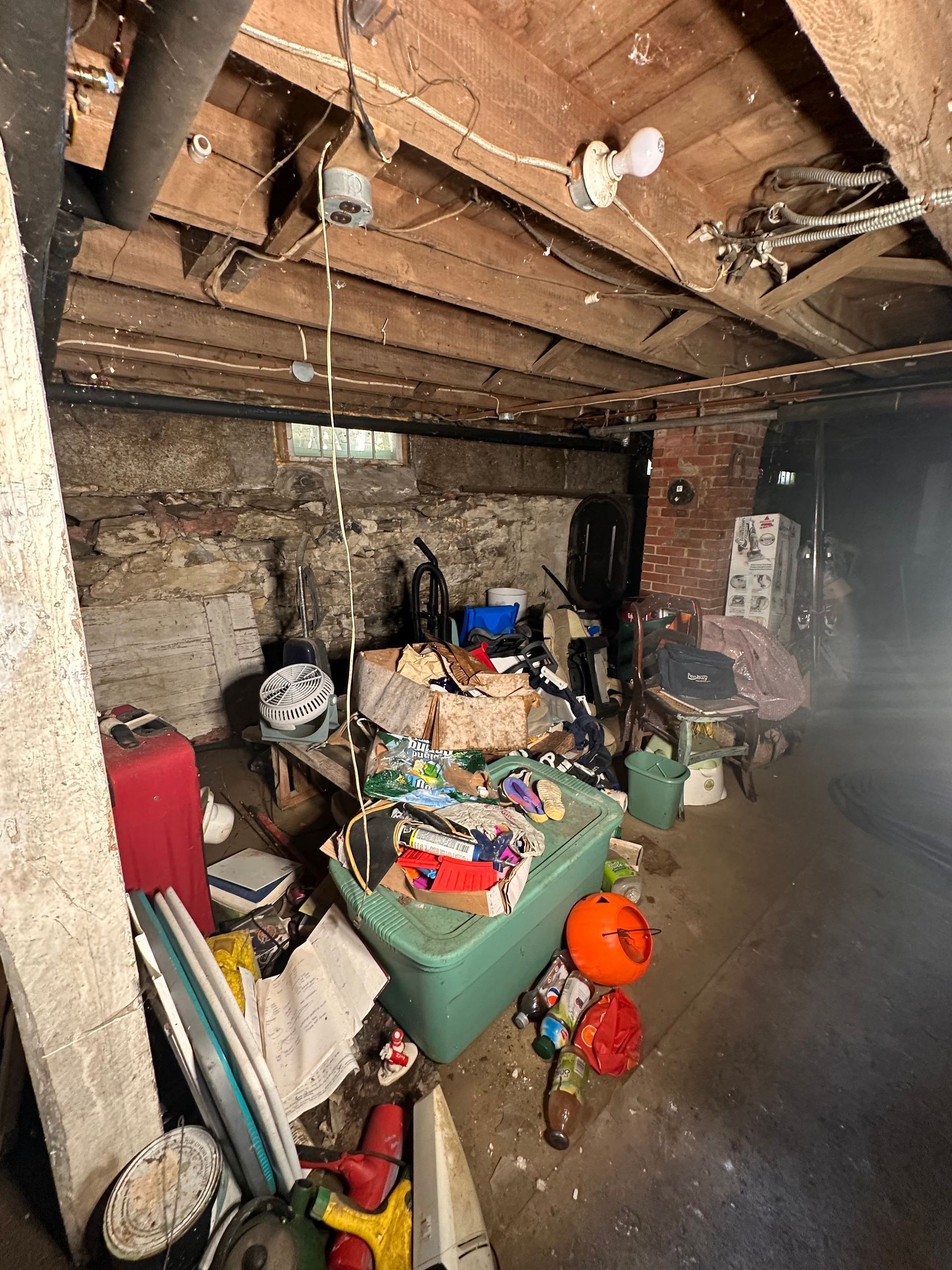 A cluttered basement with a stone wall, exposed wooden ceiling beams, a green storage bin, and various household items.