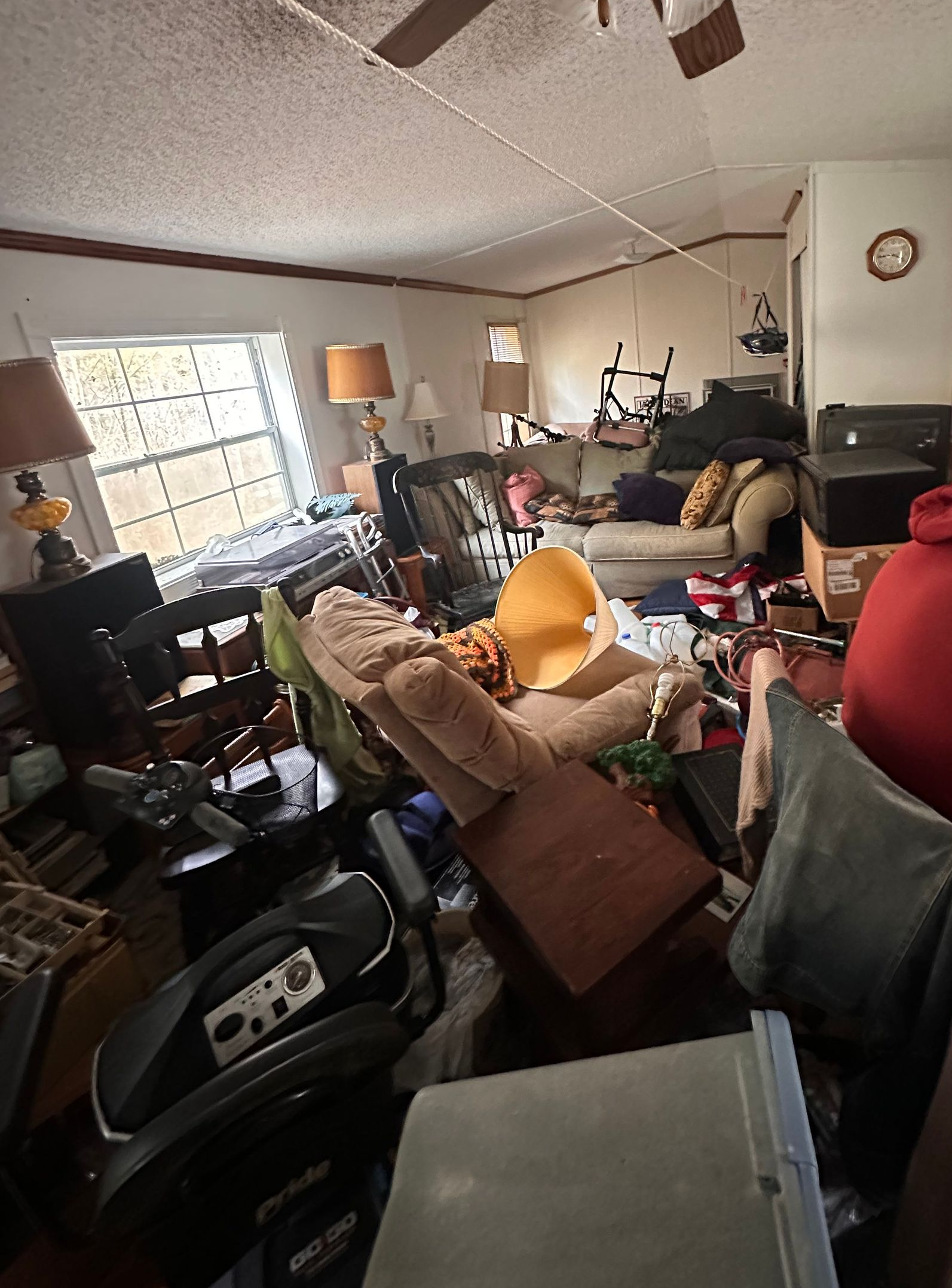 A person in a red hoodie sits in a cluttered living room filled with furniture, exercise equipment, and household items.