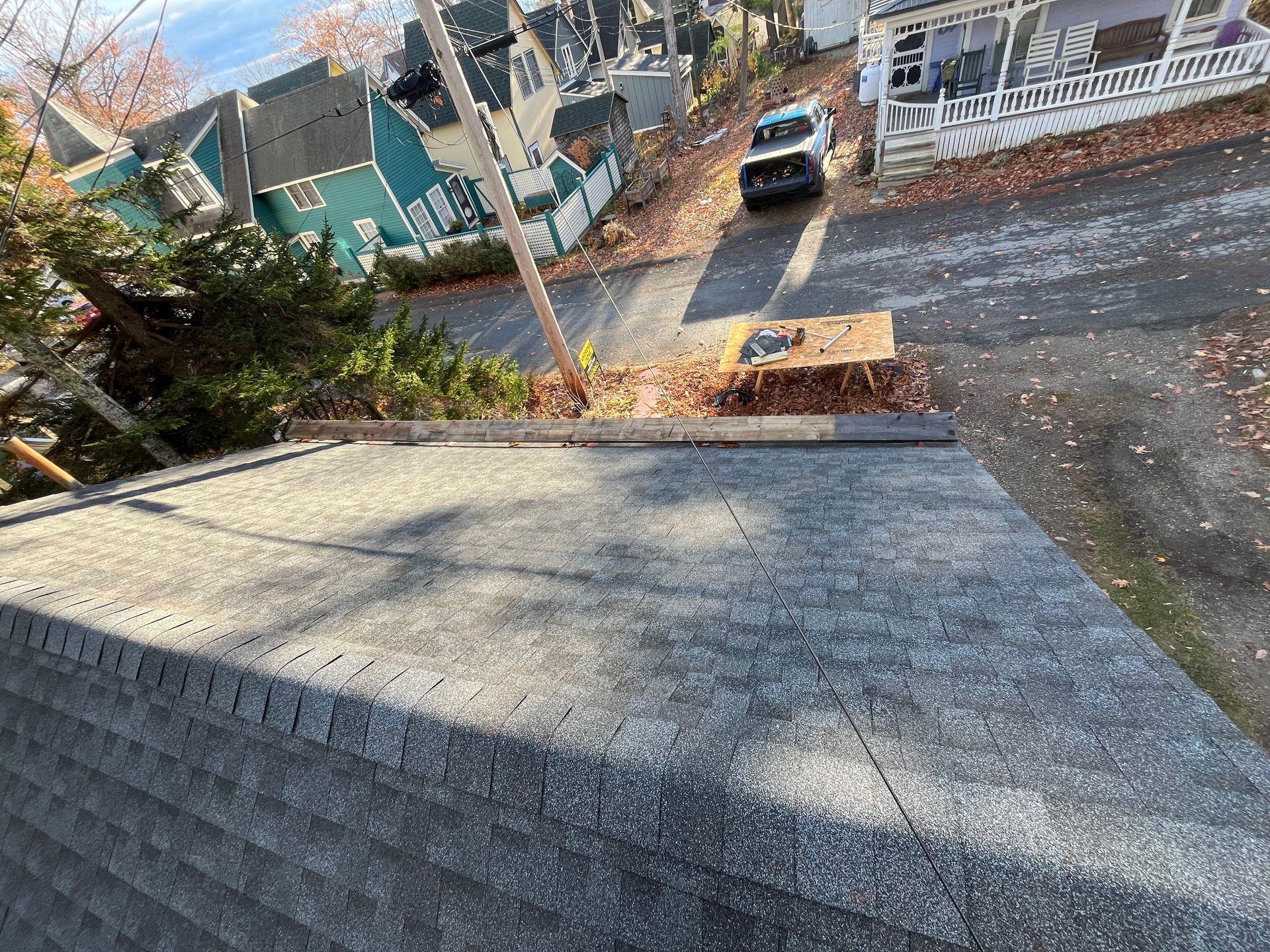 A high-angle view of a gray asphalt shingle roof, with a street, a vehicle, and houses visible in the background.