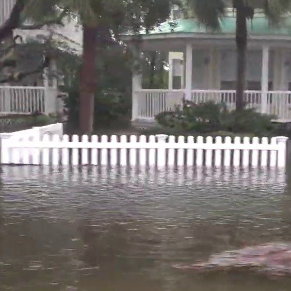 A white picket fence is surrounded by flood water in front of a house.