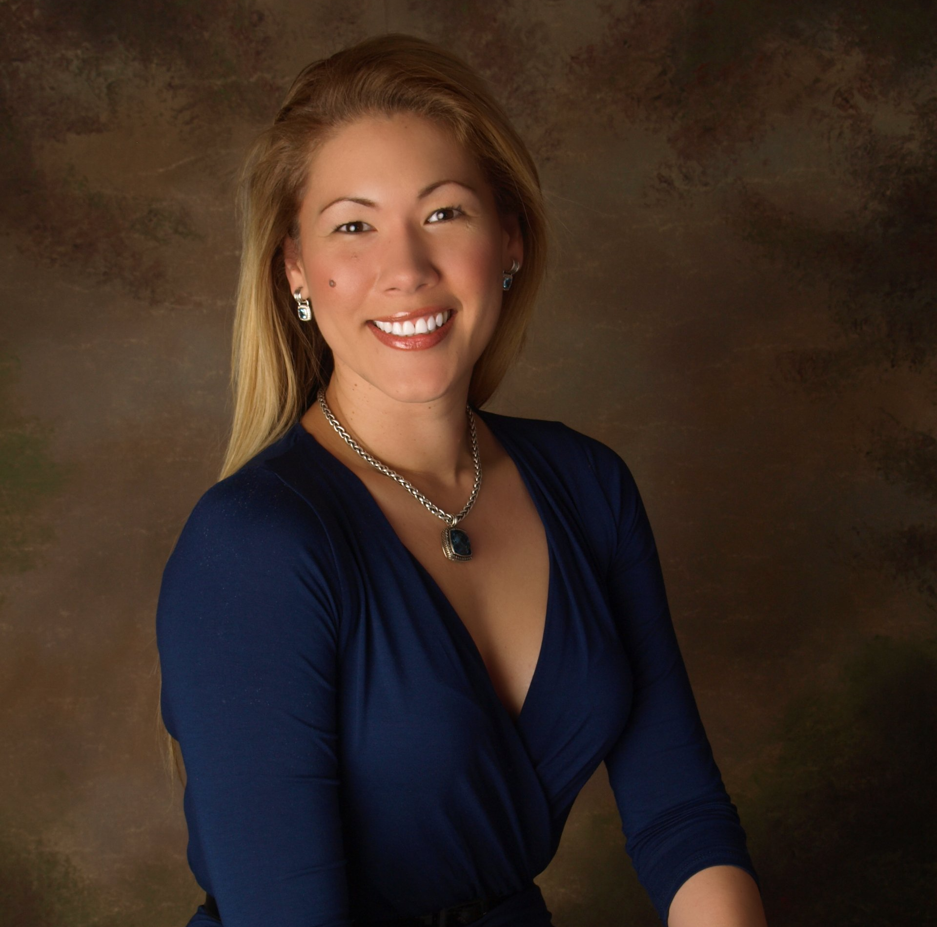 A woman wearing a blue top and a necklace smiles for the camera